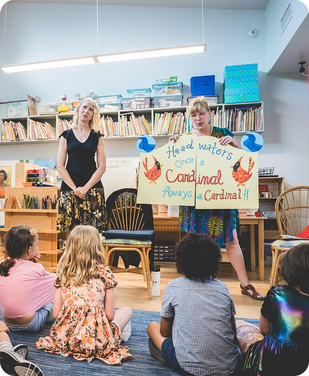 Guides hold a “Once a Cardinal, Always a Cardinal” sign while young students sit together on the classroom floor during an end-of-year gathering at Headwaters School called Gates of Life.