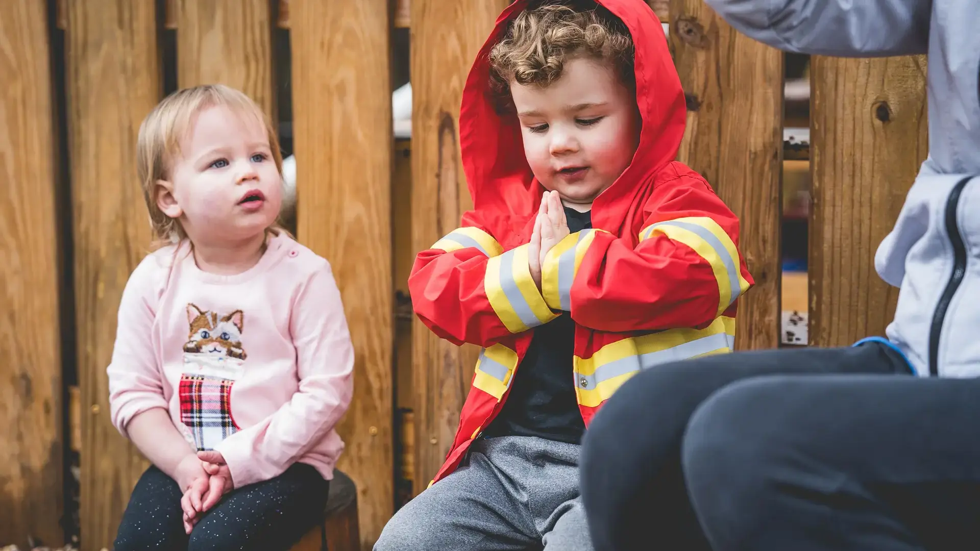 Two toddlers sitting together outdoors, one wearing a red jacket.
