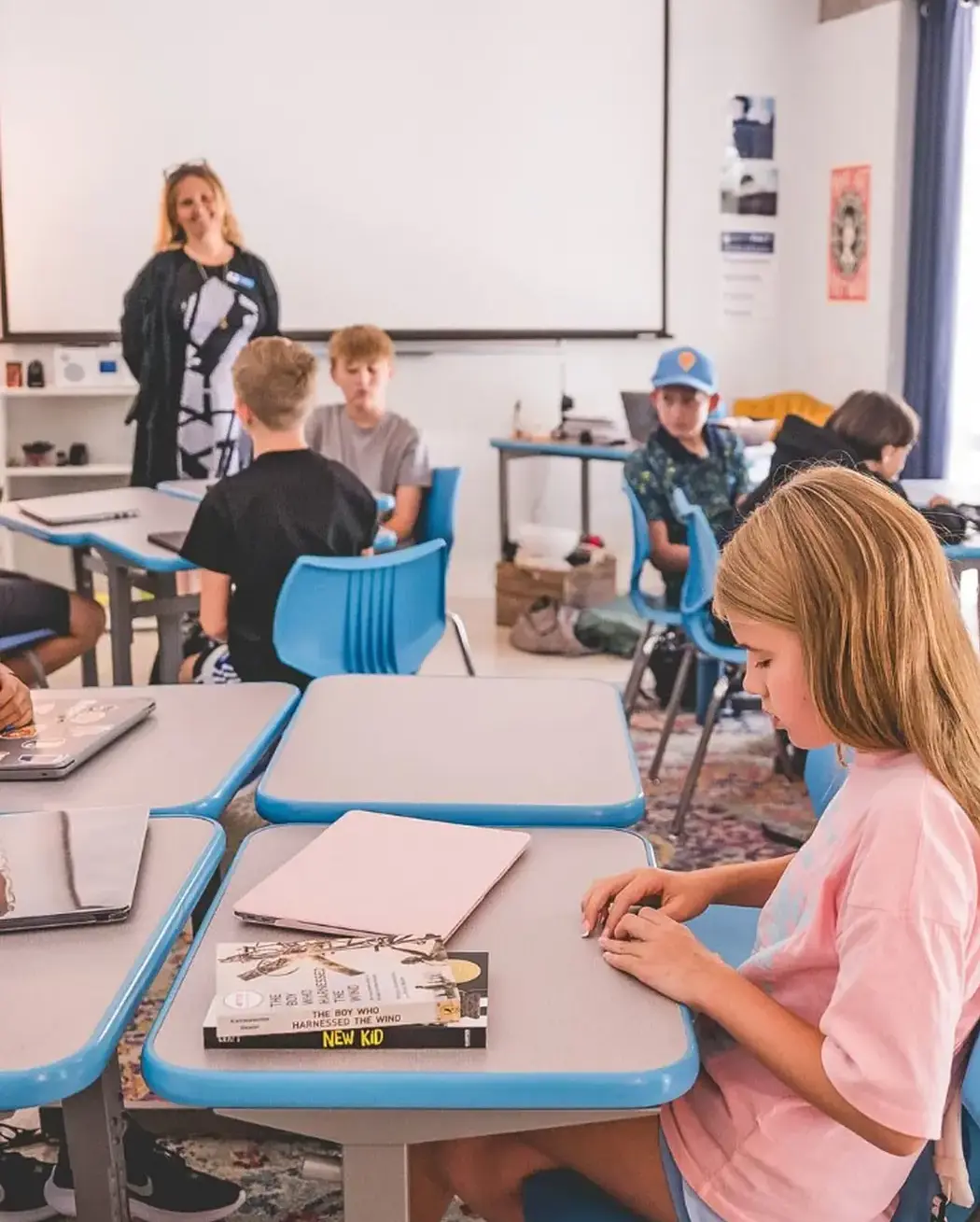 students at desks during a lesson, with a teacher standing near a projector screen at the front of the classroom.