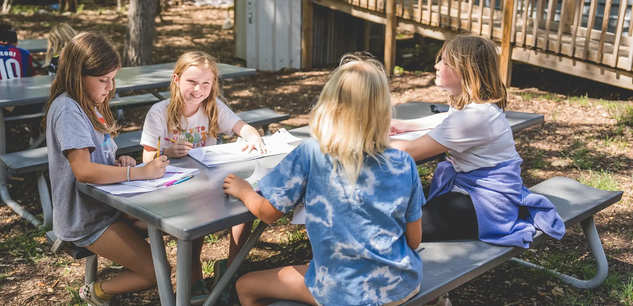 Elementary students writing and talking together at an outdoor picnic table.