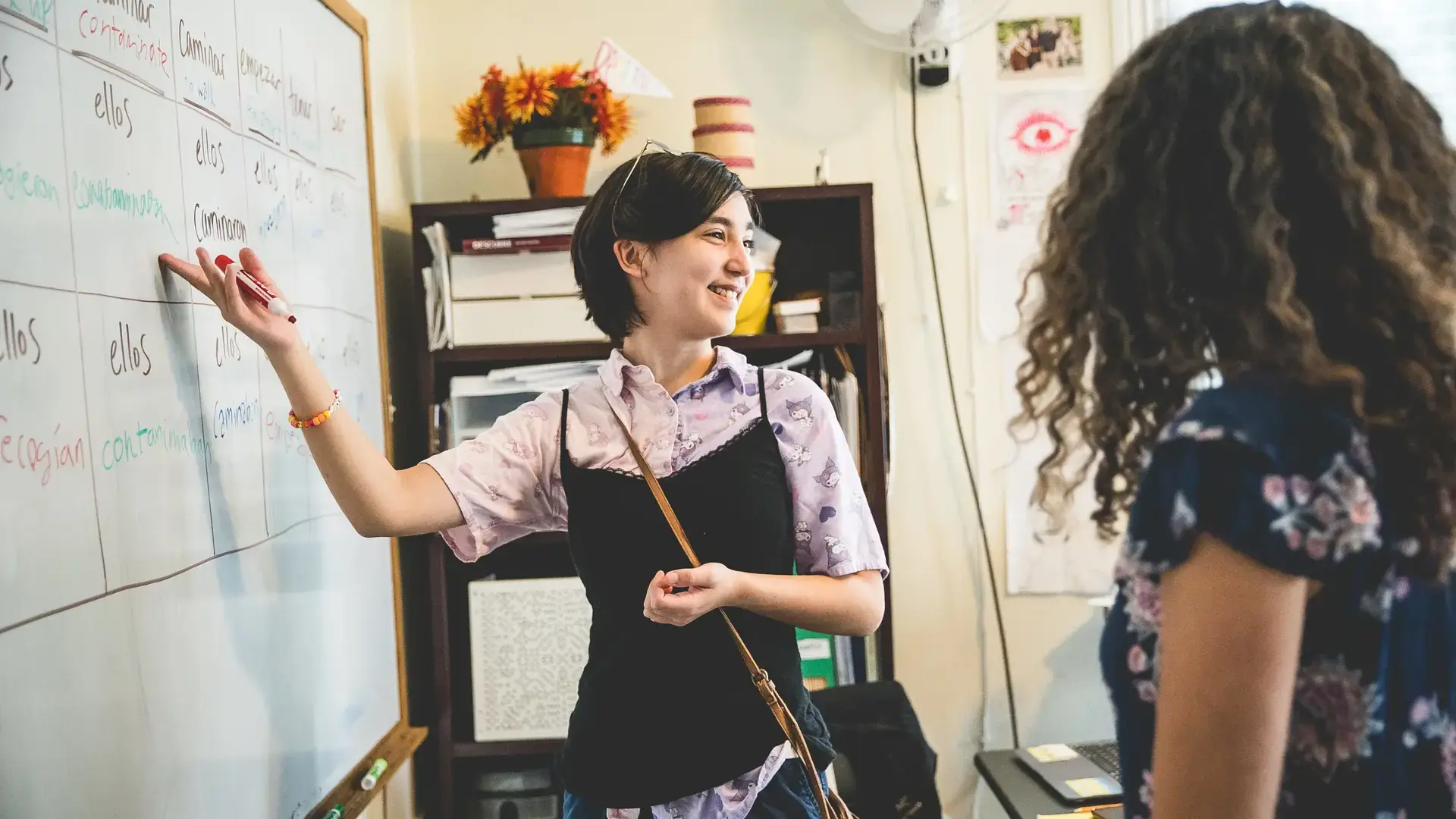 A student smiling and pointing to Spanish verb conjugations on a whiteboard while explaining to a classmate.