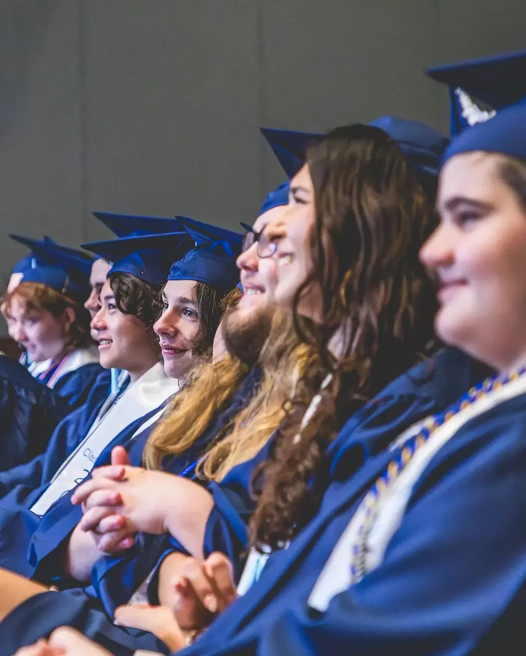 High school graduates in navy caps and gowns seated in a row, smiling during commencement.