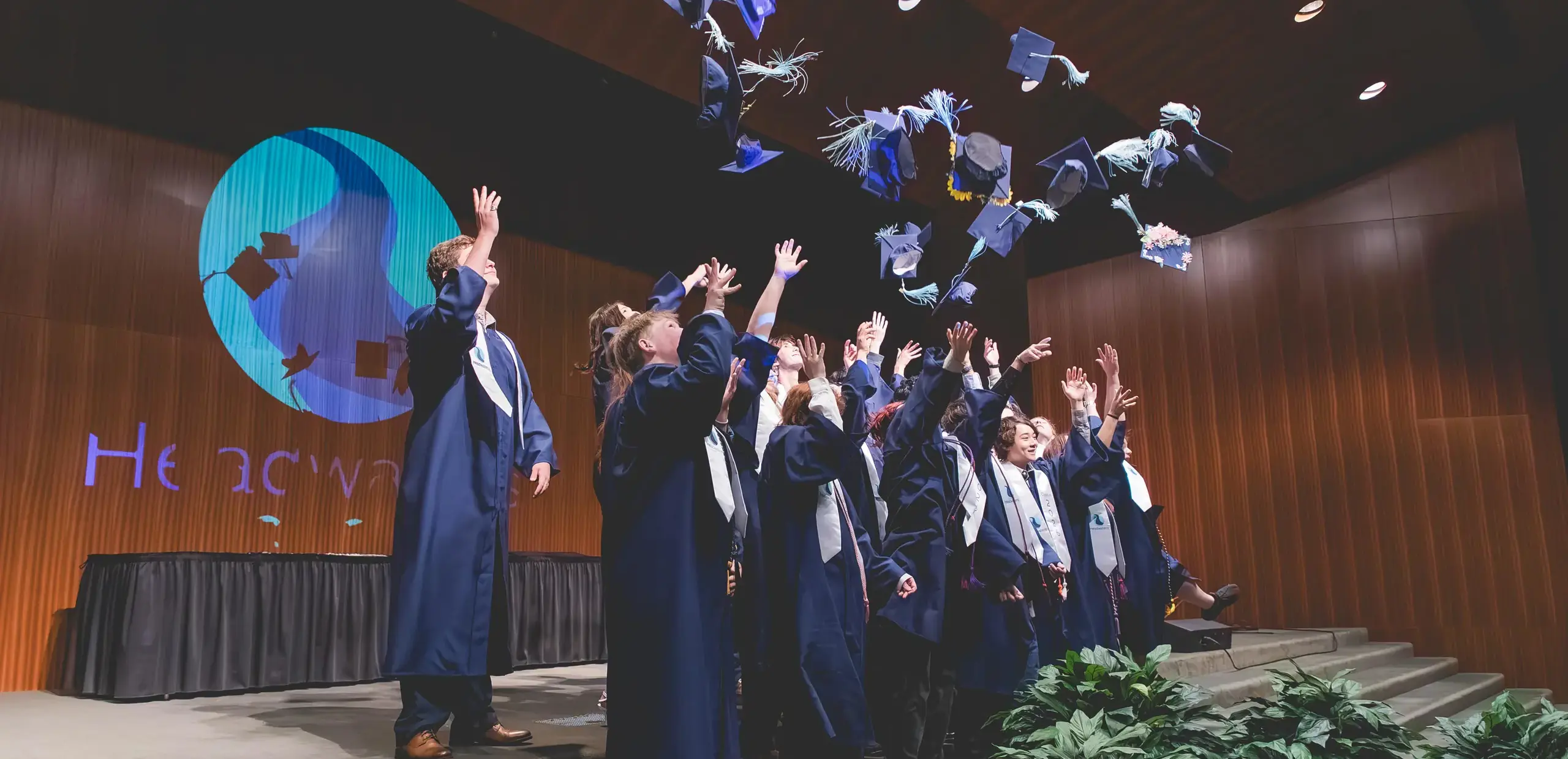Headwaters School graduates in caps and gowns tossing their decorated caps into the air on stage.
