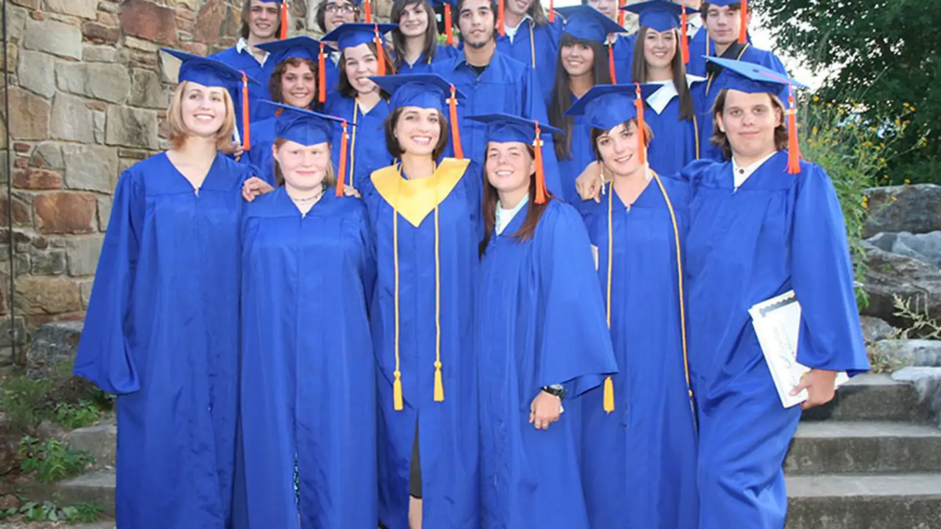 Group of graduates in royal blue caps and gowns posing together outdoors.