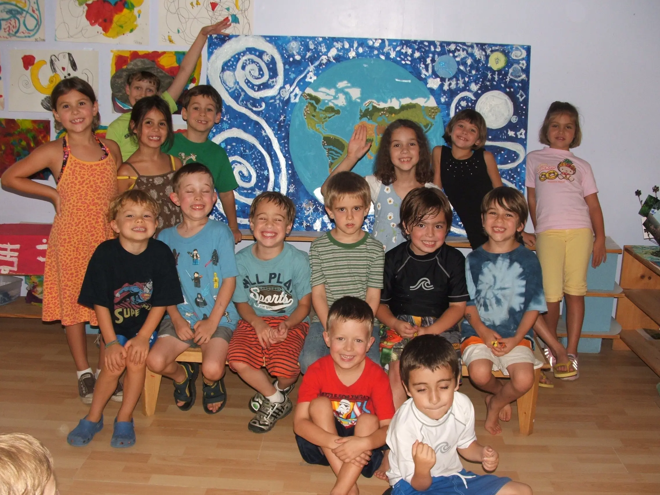Primary-aged students gathered in a classroom, posing in front of a large painted mural of the Earth and stars.