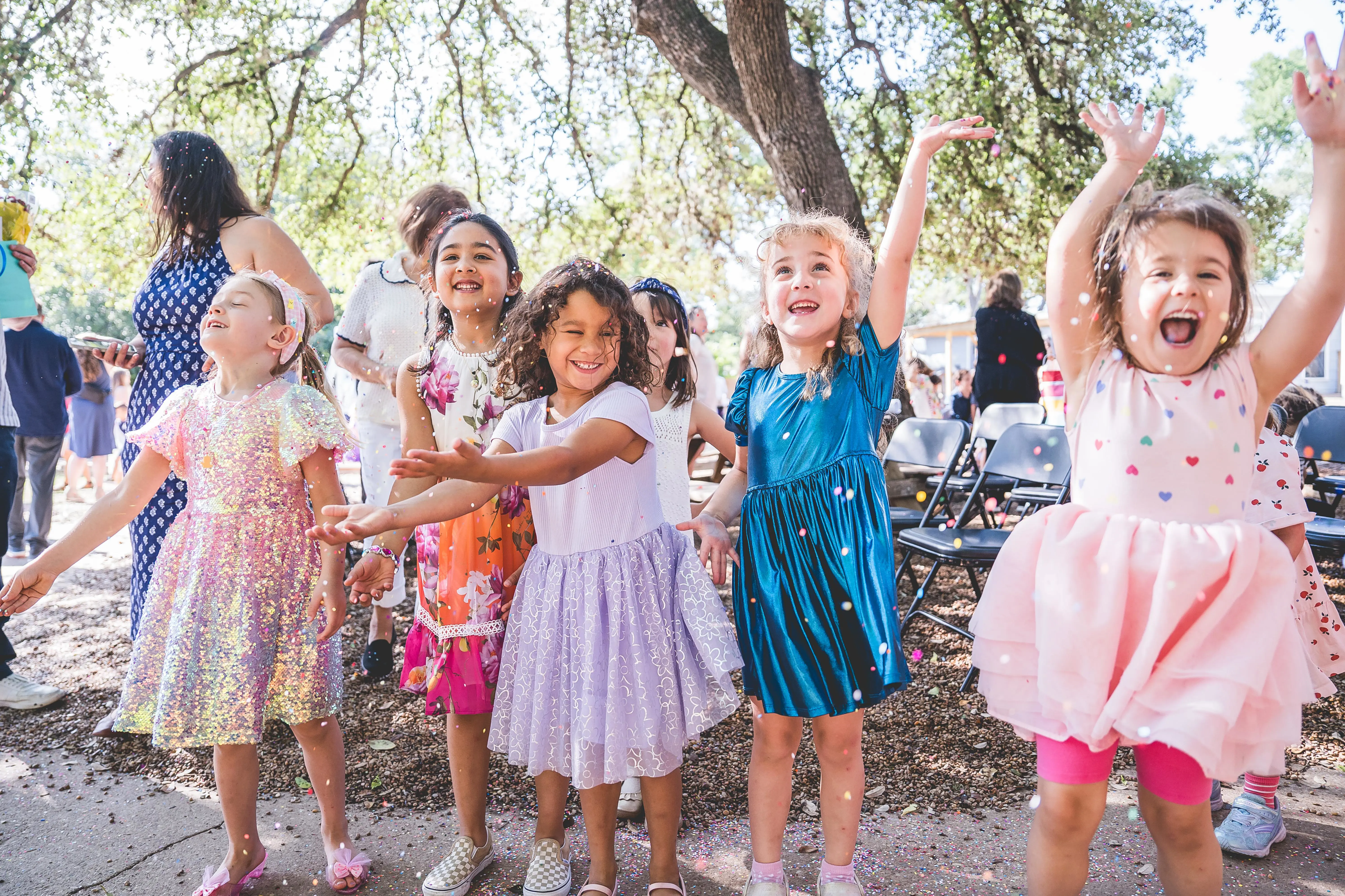 Young students in colorful dresses throw confetti and celebrate together outdoors during a Primary graduation celebration at Headwaters School.