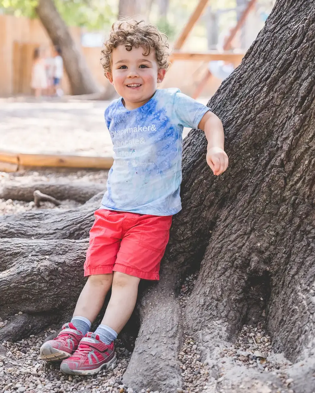 Young child smiling while leaning against a tree trunk on an outdoor playground.