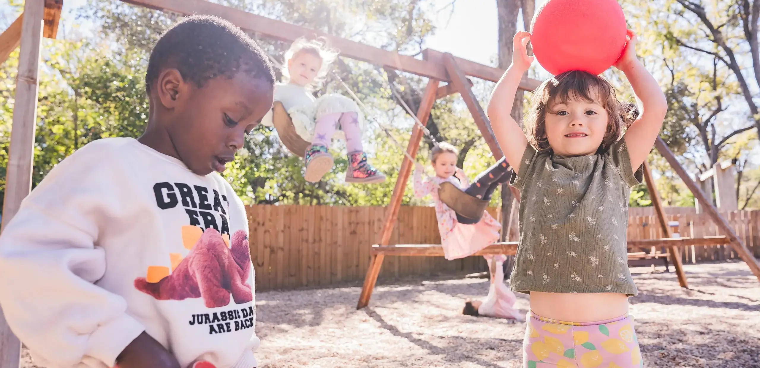 young students playing on the playground, with two swinging in the background and one holding a red ball overhead.