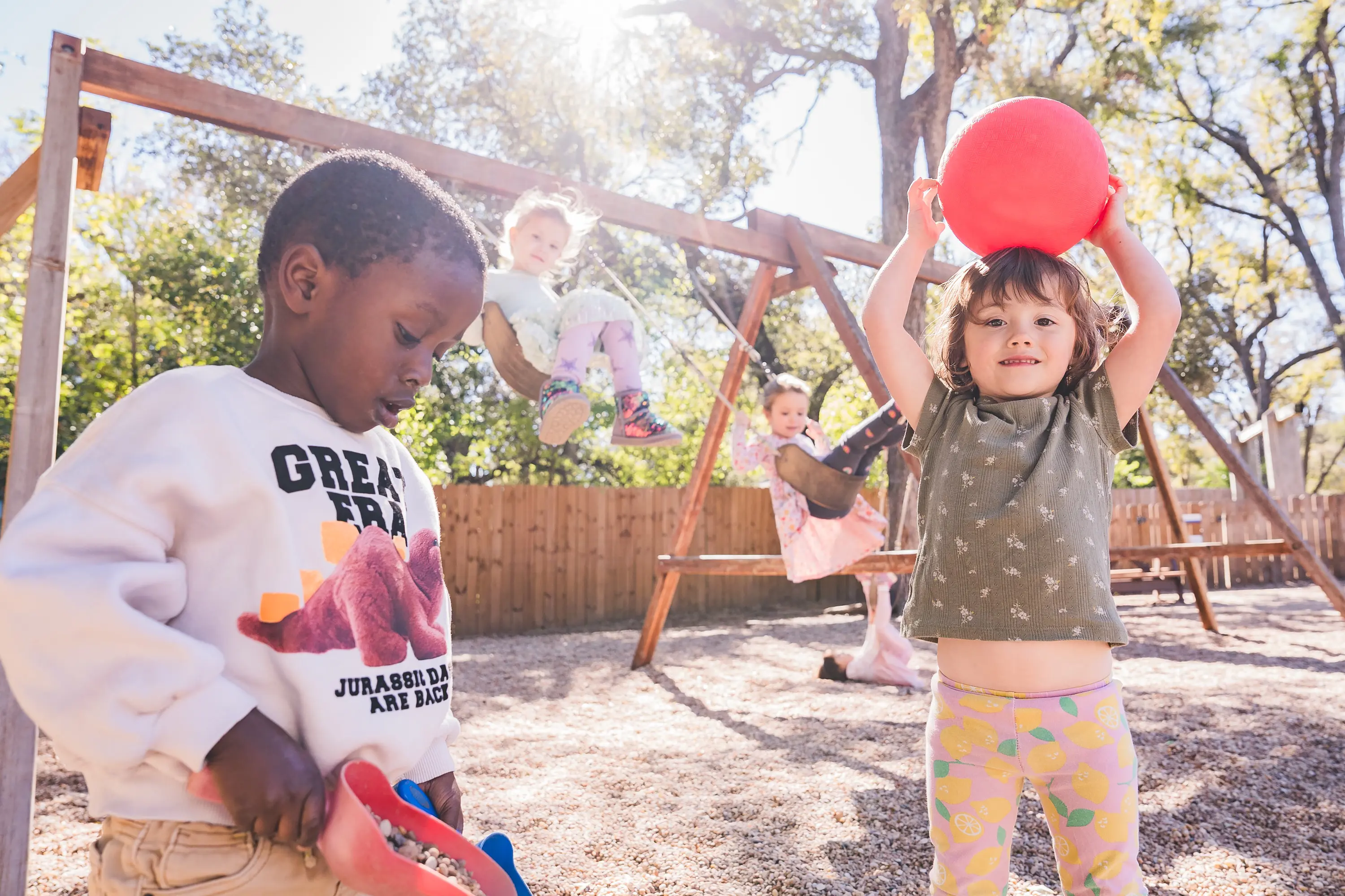 Four young children playing outdoors on a playground, with two children on swings, one child holding a red ball overhead, and another scooping gravel.