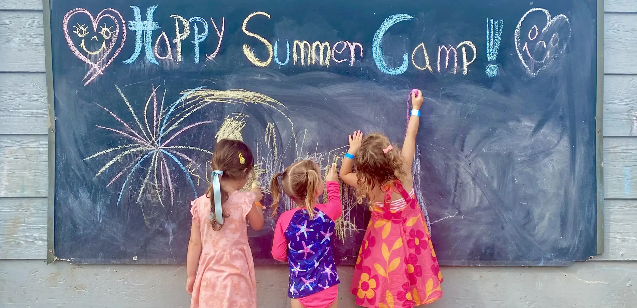 Three young children drawing fireworks and 'Happy Summer Camp' on an outdoor chalkboard.