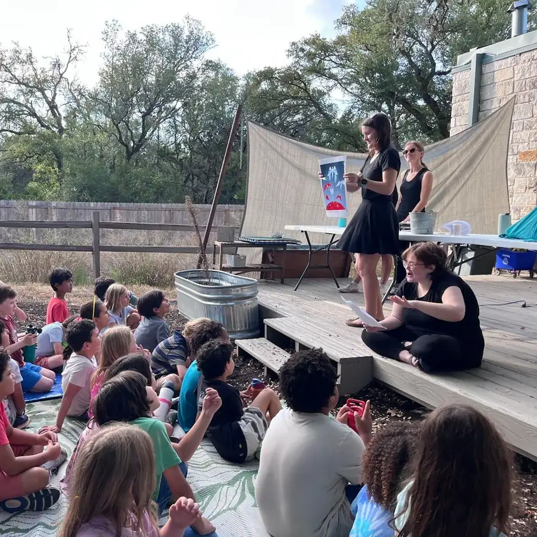 Early Elementary students sit outdoors as guides present the First Great Lesson, exploring the Big Bang through storytelling and science demonstrations in a shared courtyard setting.