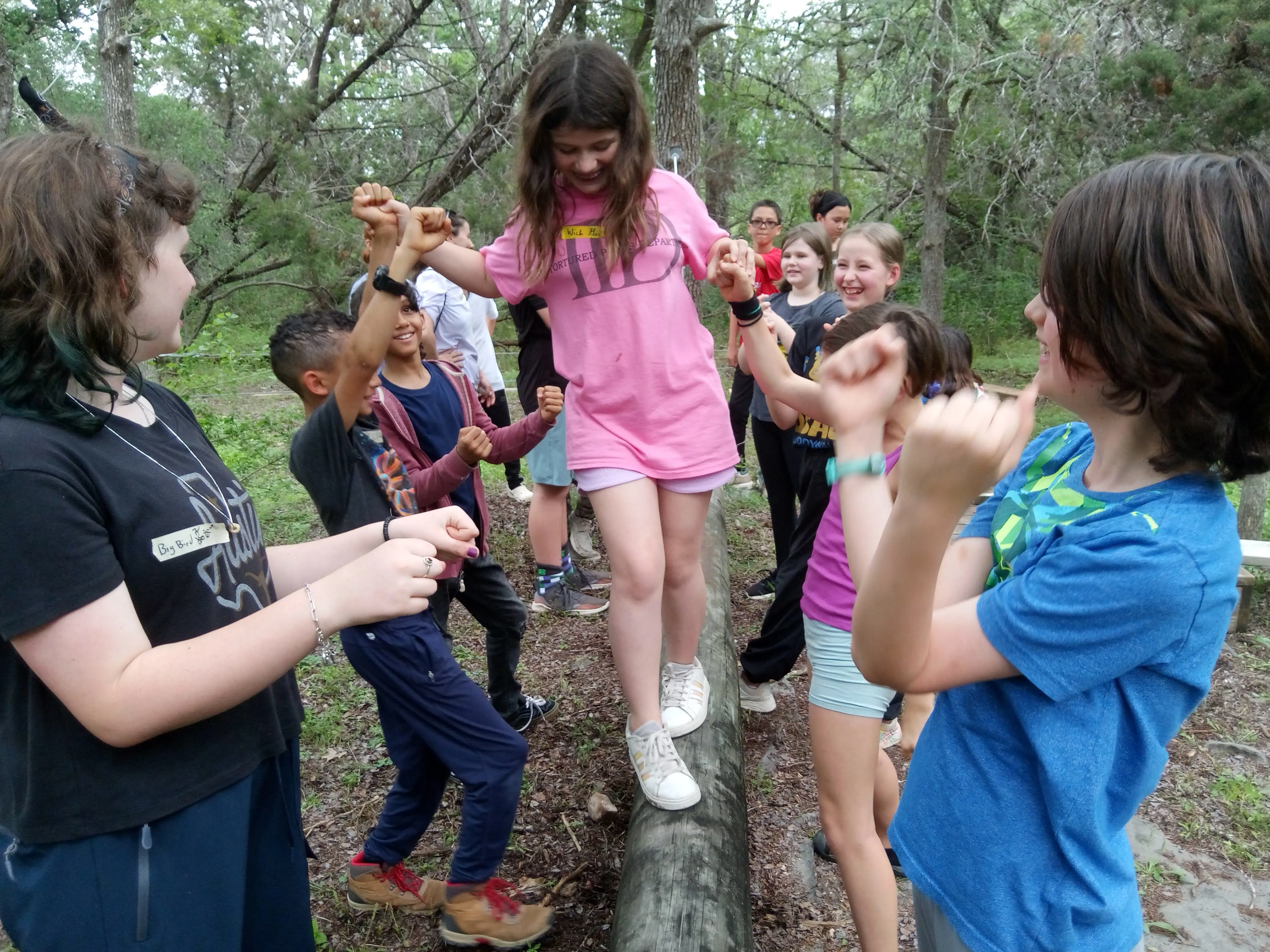 Elementary students gather in the Bird’s Nest outdoor learning area, helping a classmate balance across a log as they cheer and support one another during a group challenge in the woods.