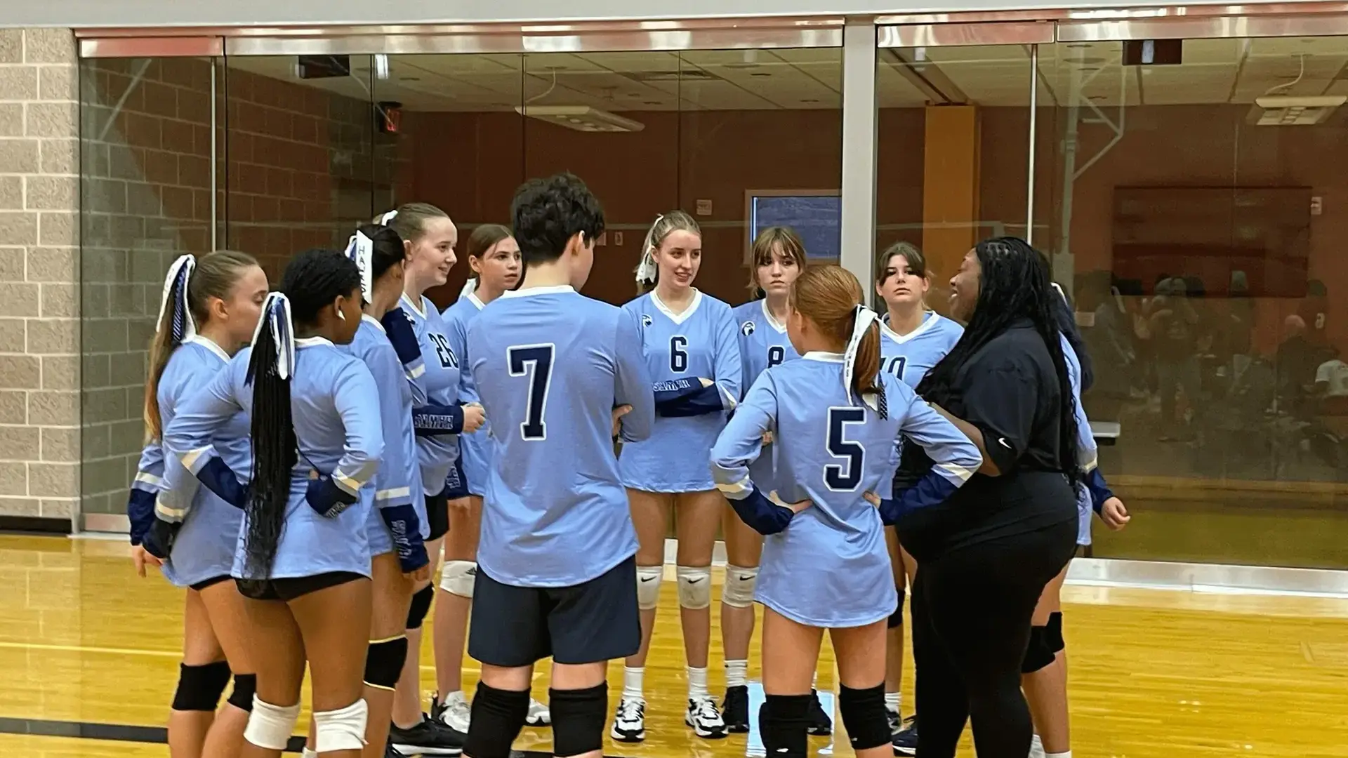 A girls volleyball team huddled together in matching light blue jerseys during a game.