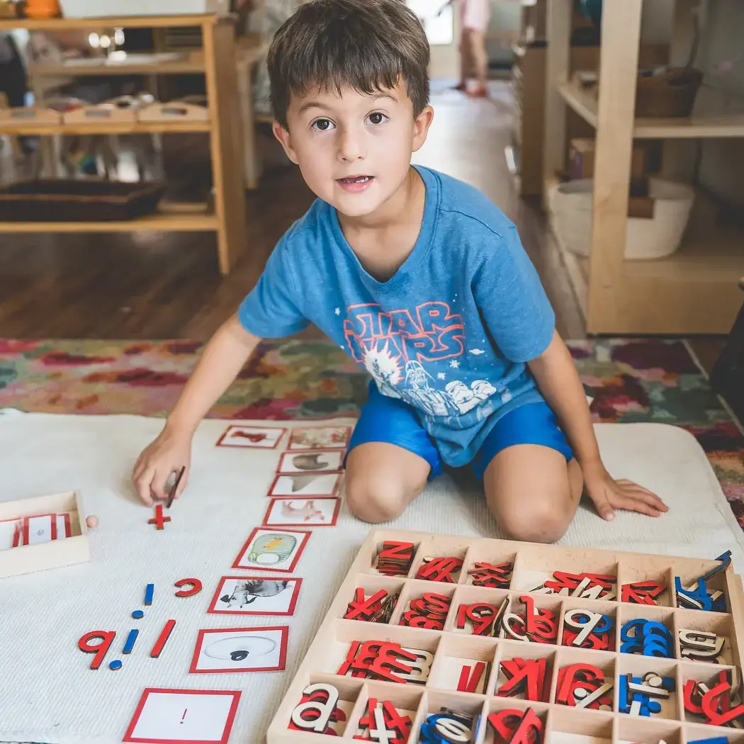 Primary student works on a Montessori language activity with moveable alphabet letters and picture cards while seated on a classroom floor mat.