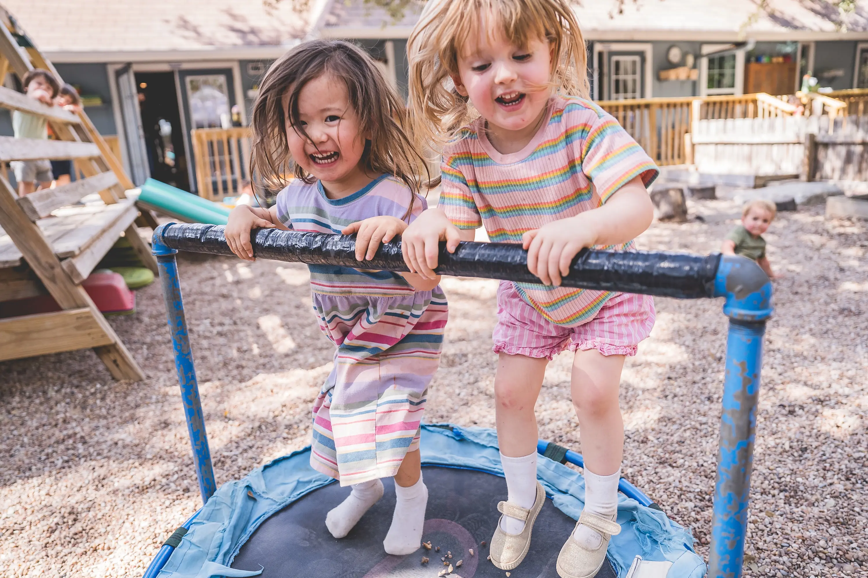 Two young children laugh while bouncing on a small trampoline during outdoor play at the Headwaters Young Children’s Community playground.