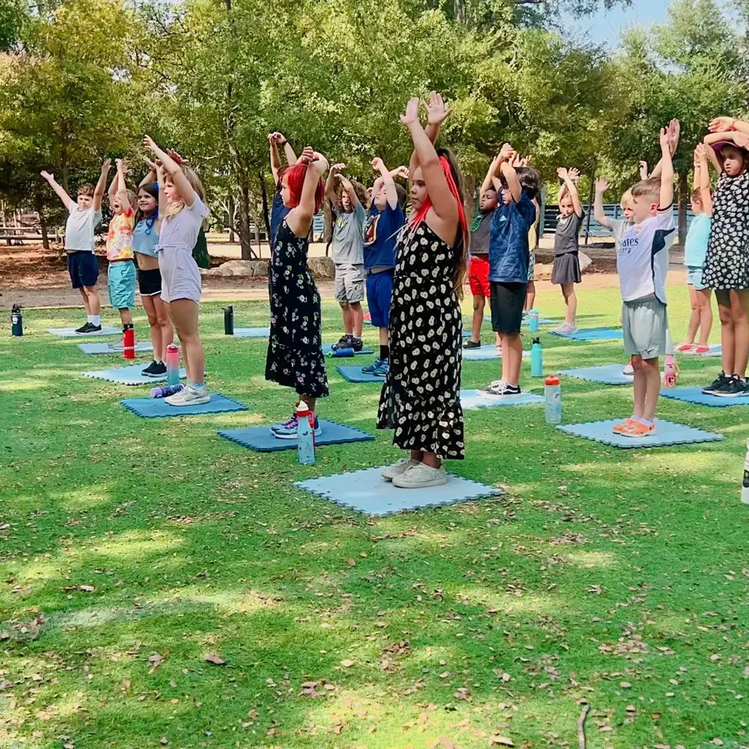 elementary students standing on mats outdoors with arms raised during a yoga class.