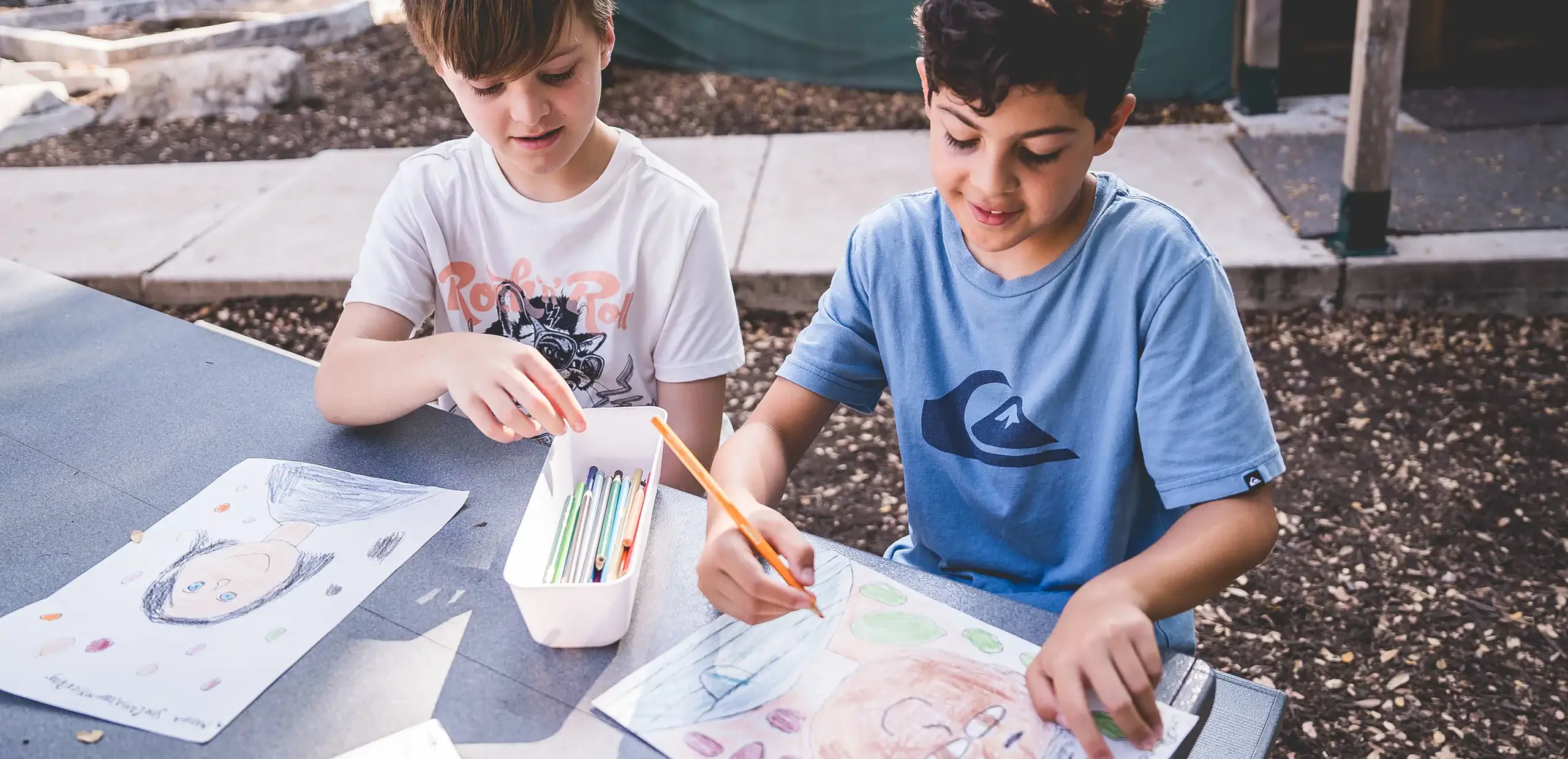 two elementary students drawing self-portraits together at an outdoor table.