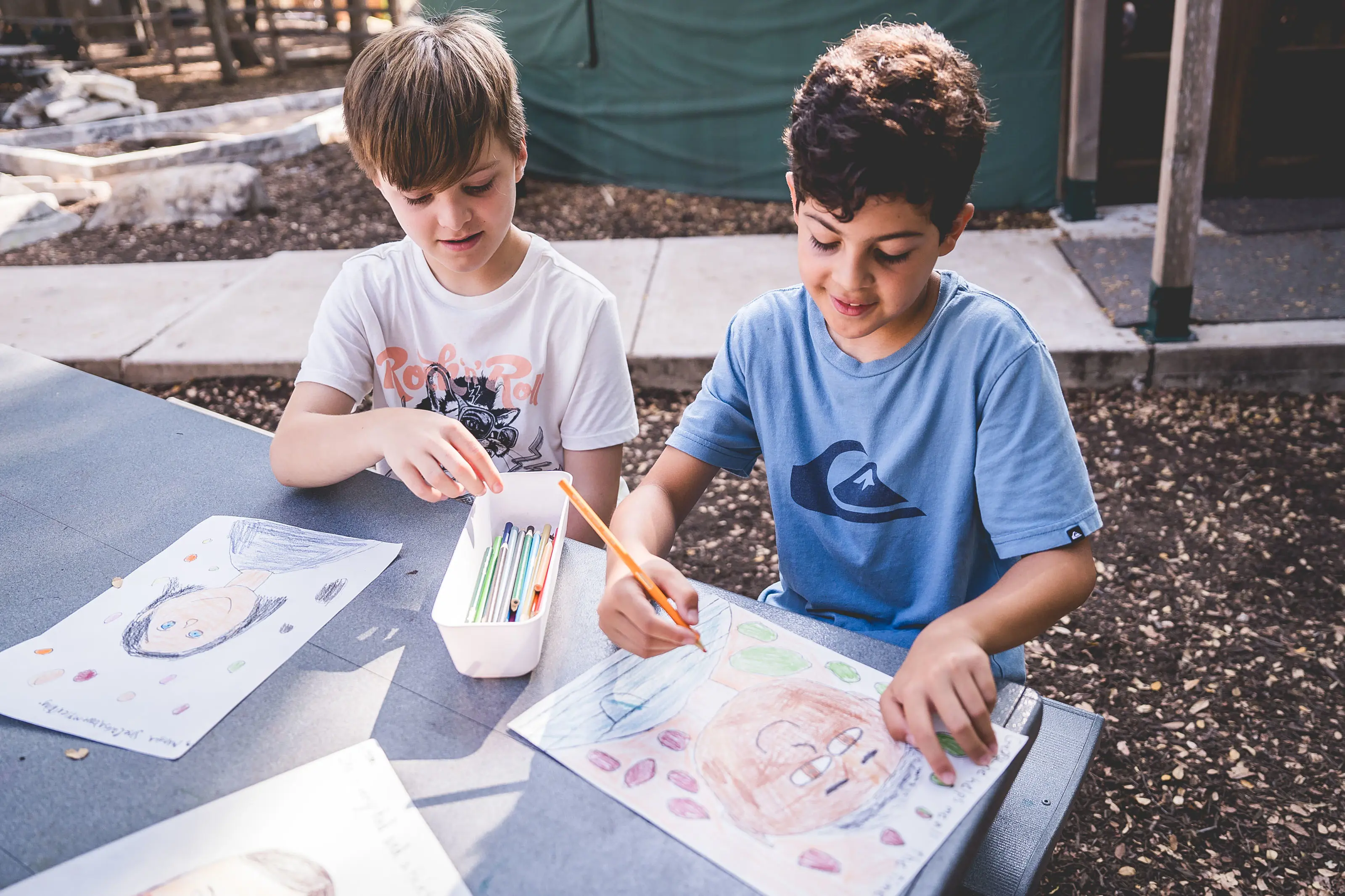 Two elementary students sit at an outdoor table drawing colorful self-portraits with colored pencils during art class.