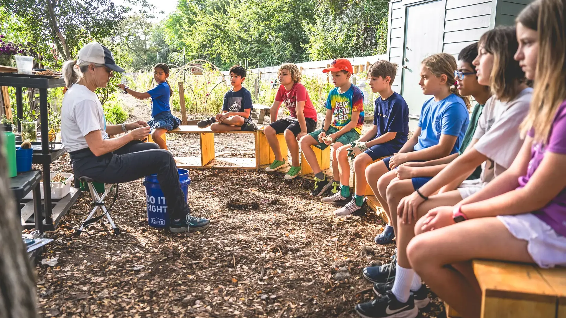 Elementary students gathered outdoors as a teacher leads a gardening lesson in the school garden.