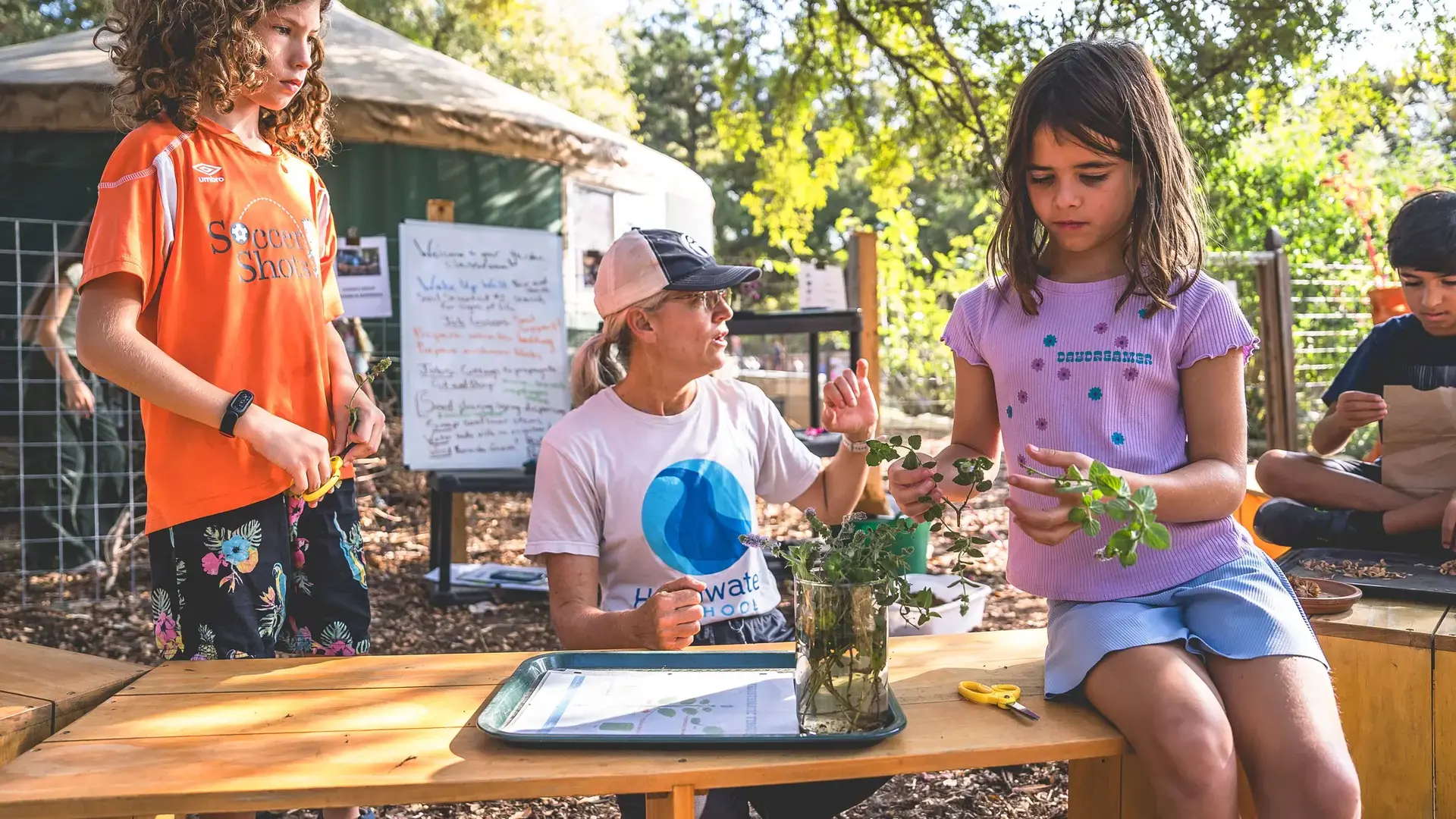 Elementary students working with a gardening teacher outdoors at a table with plants
