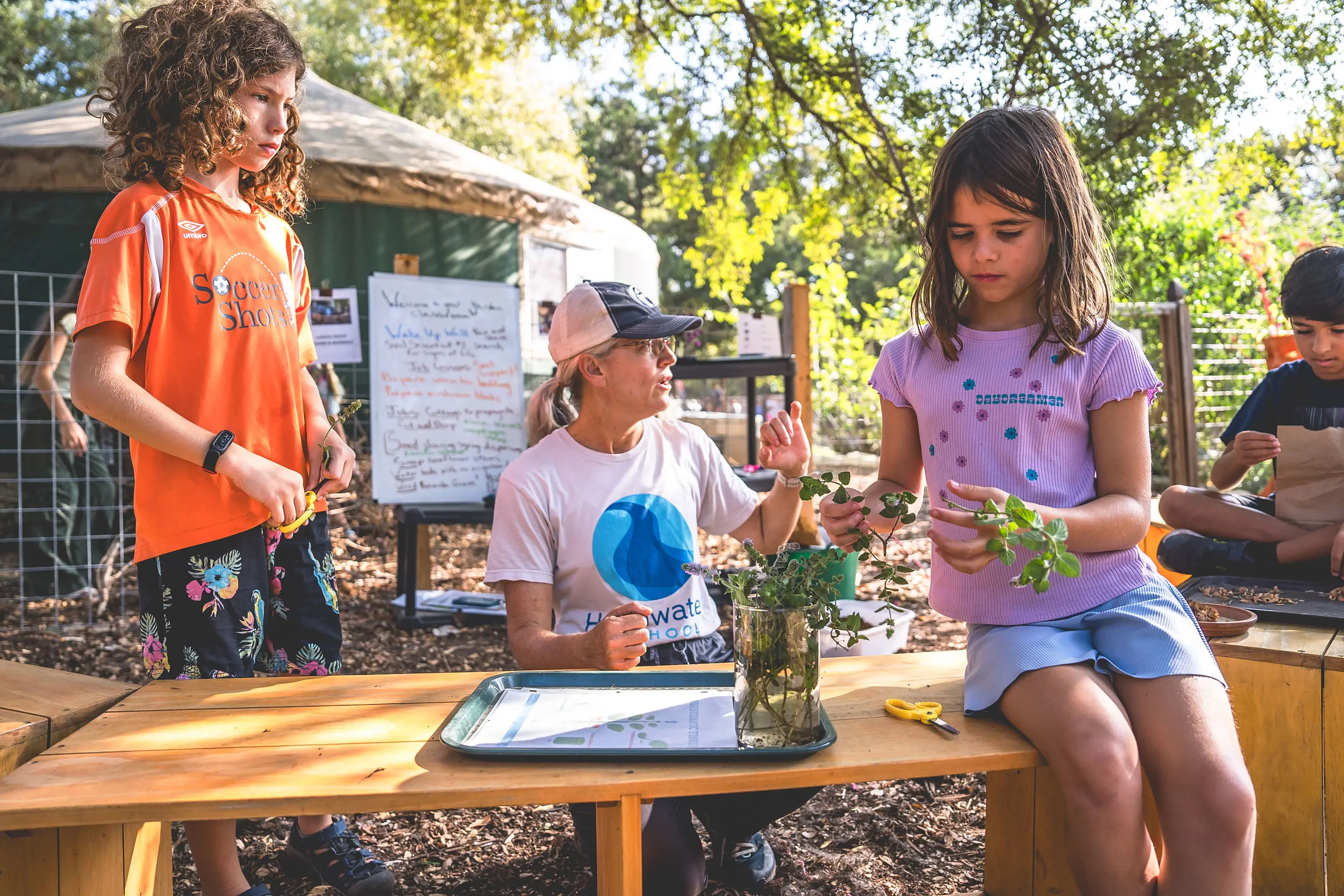 An elementary teacher guides students examining plant cuttings during a hands-on science lesson in an outdoor classroom garden at Headwaters School.