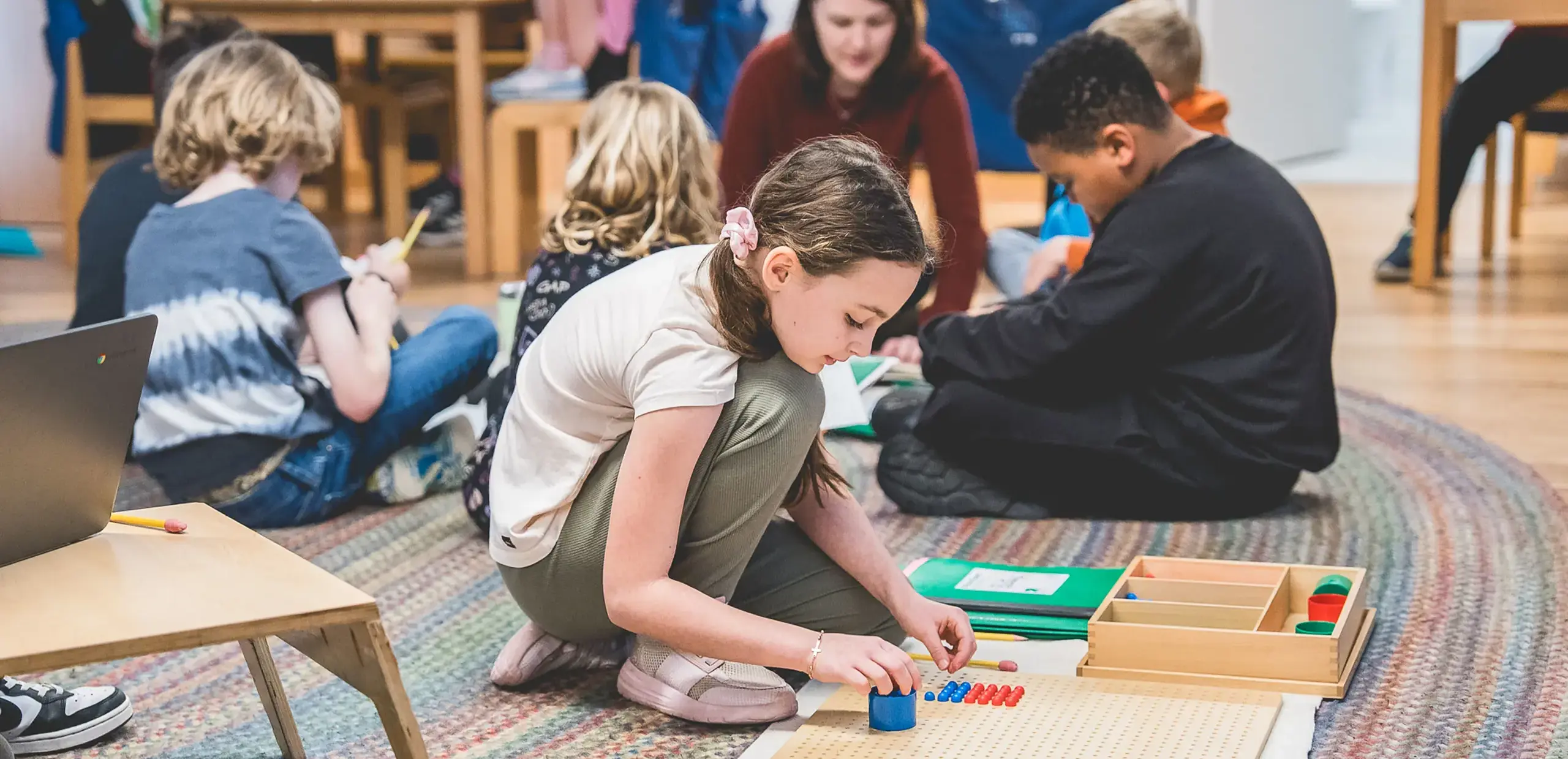 elementary students working on the floor with a teacher, using a Montessori bead board for a math activity.