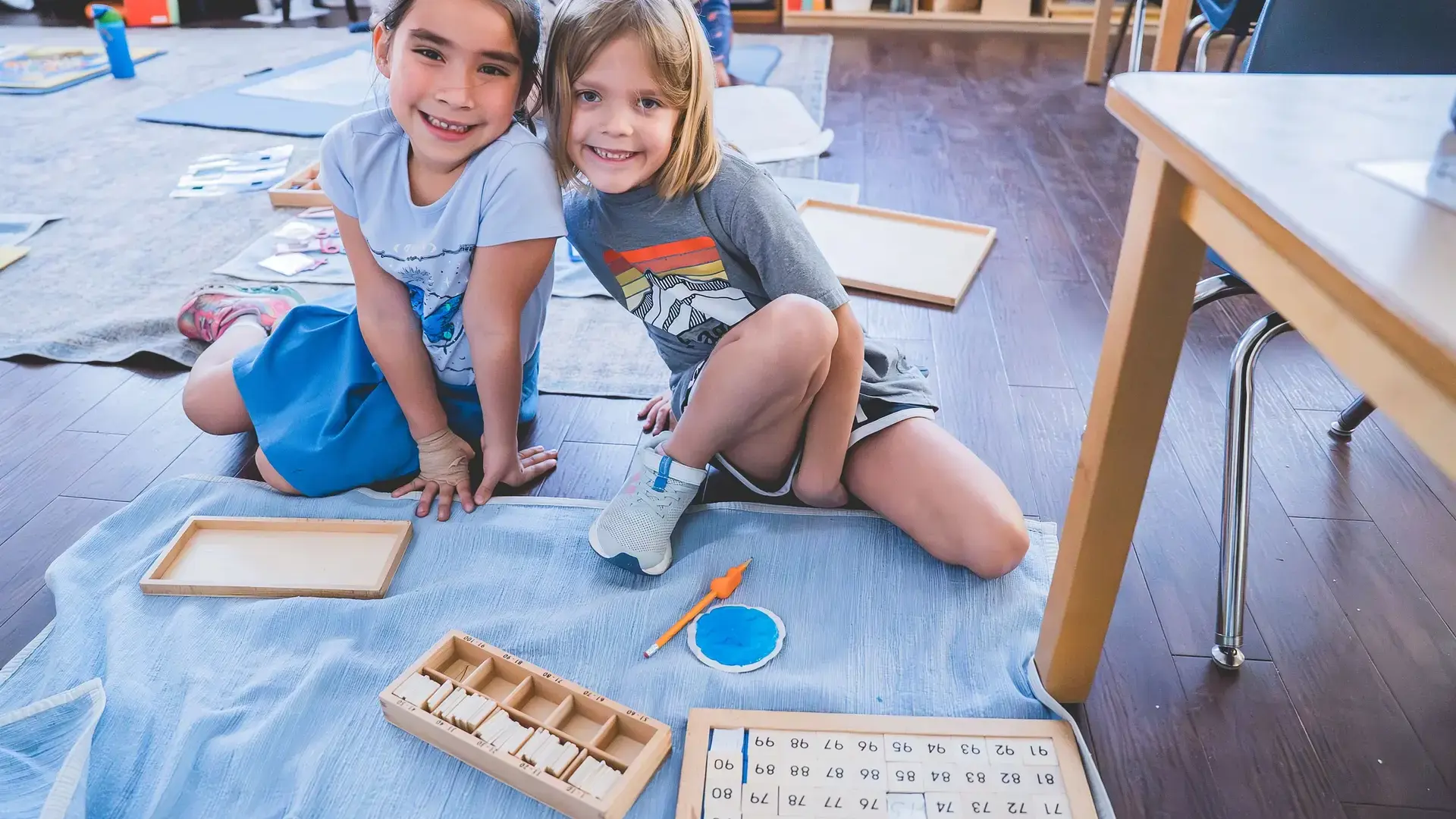 elementary students smiling while working with a Montessori math material on the floor.