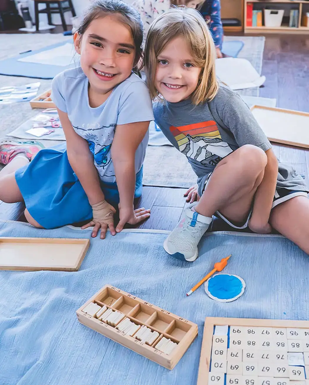 Two elementary students smiling together while working with Montessori math materials on the classroom floor.