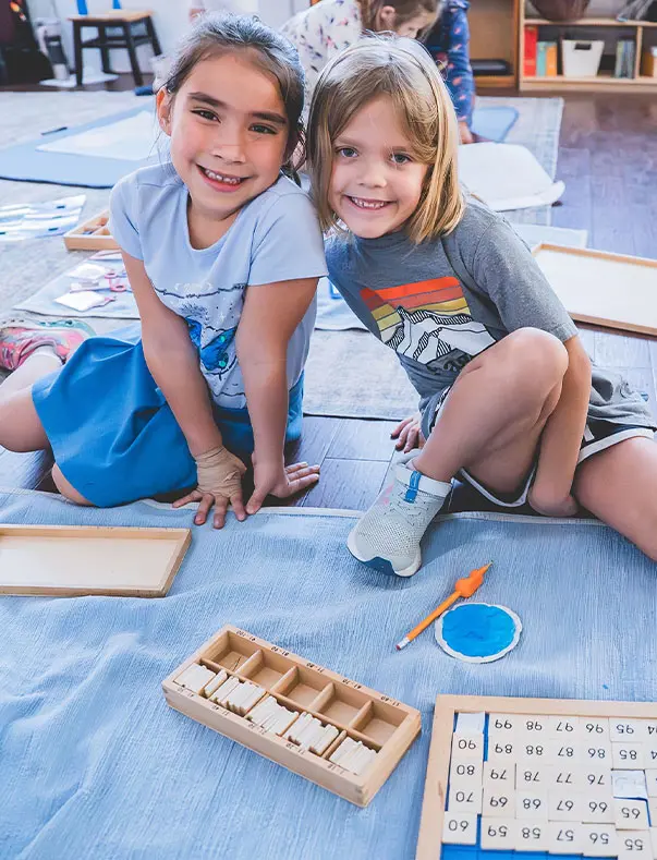 Two first graders working together on Montessori math with a work mat.  High schooler working with a calculator on a math problem