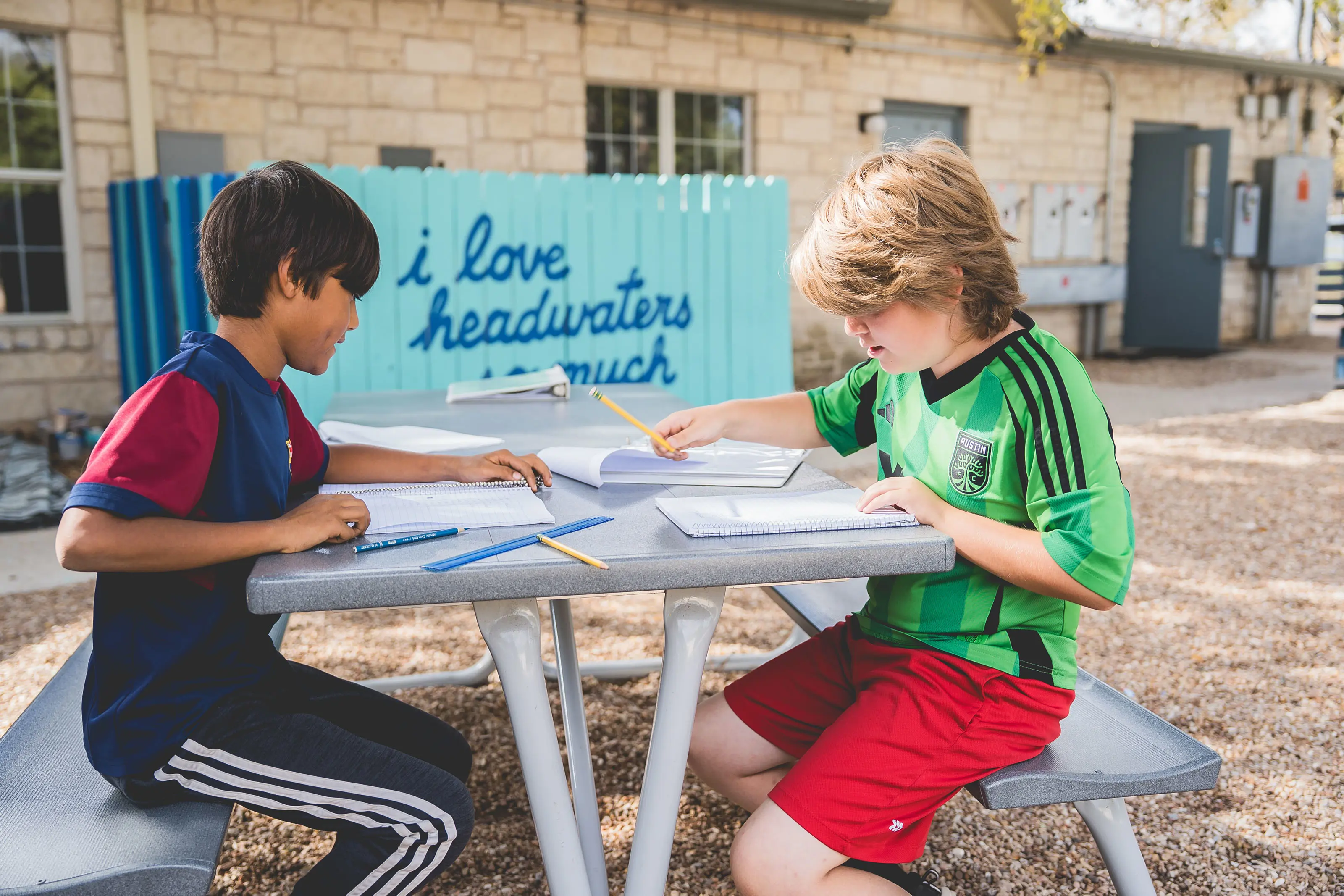 Two elementary-aged students sit at an outdoor picnic table on a school campus, working together in notebooks with pencils in hand. A turquoise fence behind them features the phrase “i love headwaters