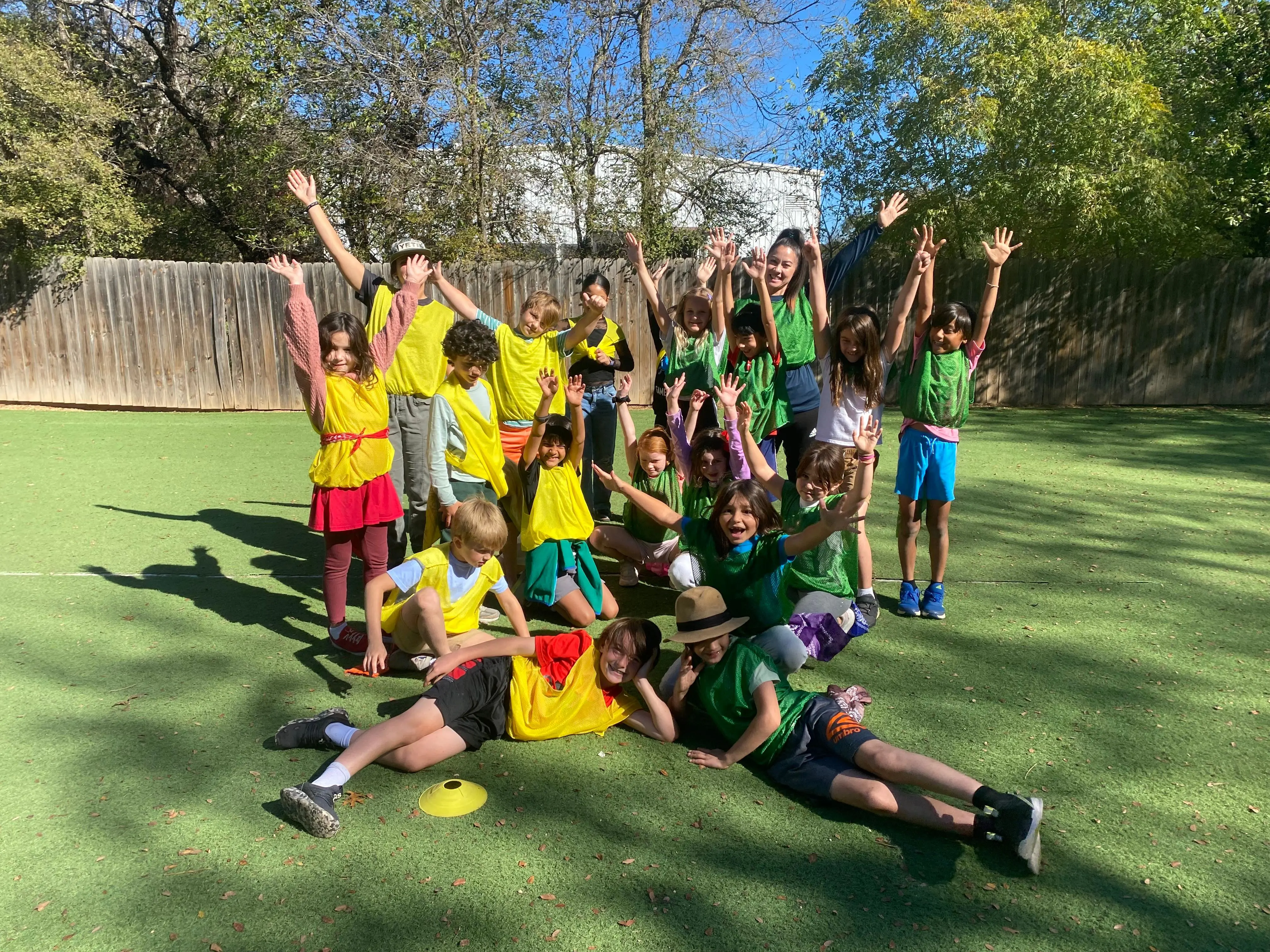 Elementary students wearing green and yellow pinnies pose together on a field after a soccer game, smiling and raising their arms in celebration outdoors on campus.
