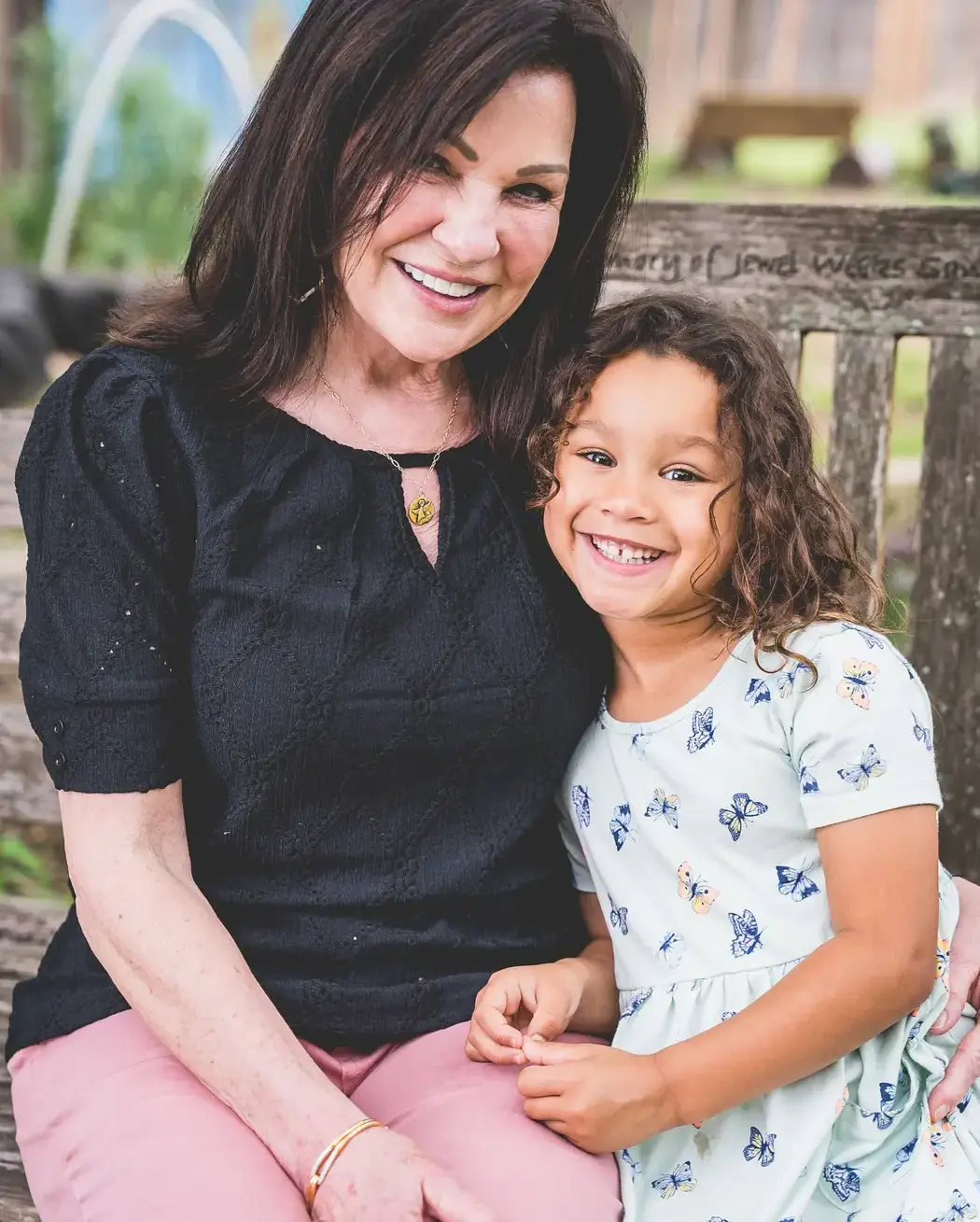 parent and a young girl smiling together while sitting on an outdoor bench.