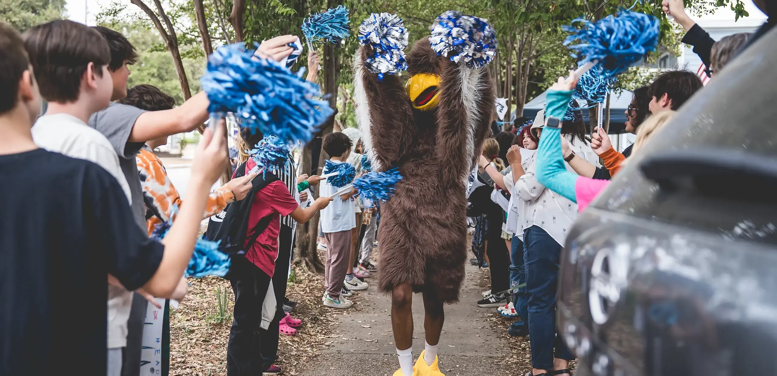 Headwaters School hawk mascot walks through a cheering crowd of students waving blue pom-poms during a school spirit celebration.