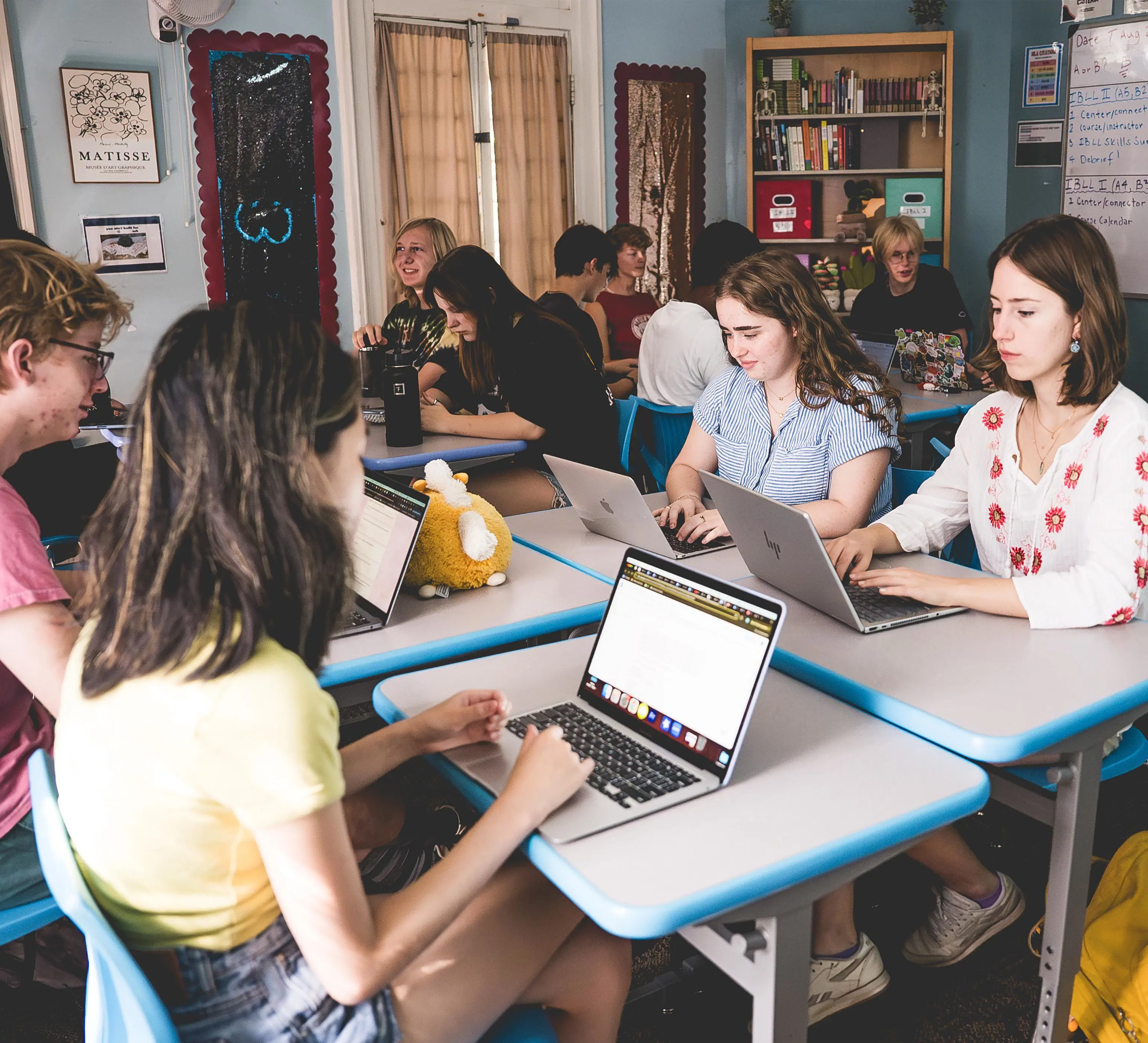 Students work on laptops at desks in a classroom decorated with art posters and bookshelves.