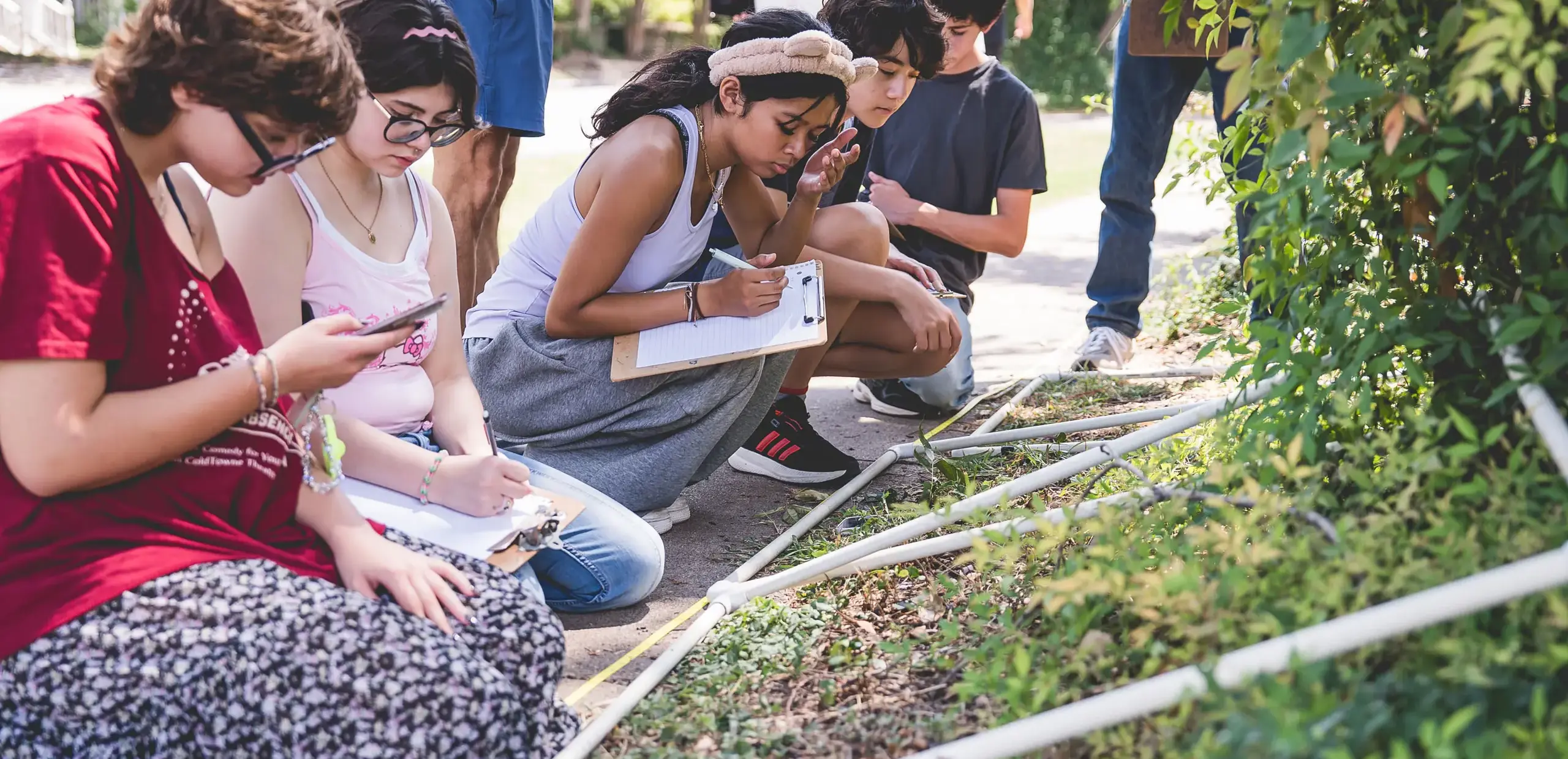 high school students recording observations on clipboards during an outdoor field science study near a garden plot.