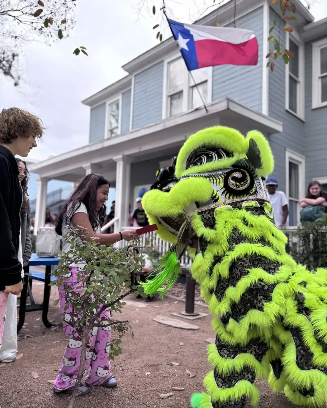 Students watching a green lion dance costume during an outdoor cultural celebration, with a Texas flag flying above.