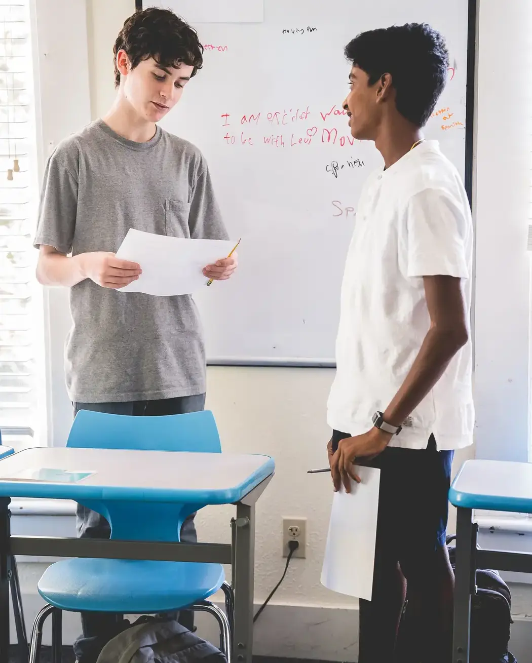 Two students talk in front of a whiteboard with student responses to "What are you excited about in this class?"