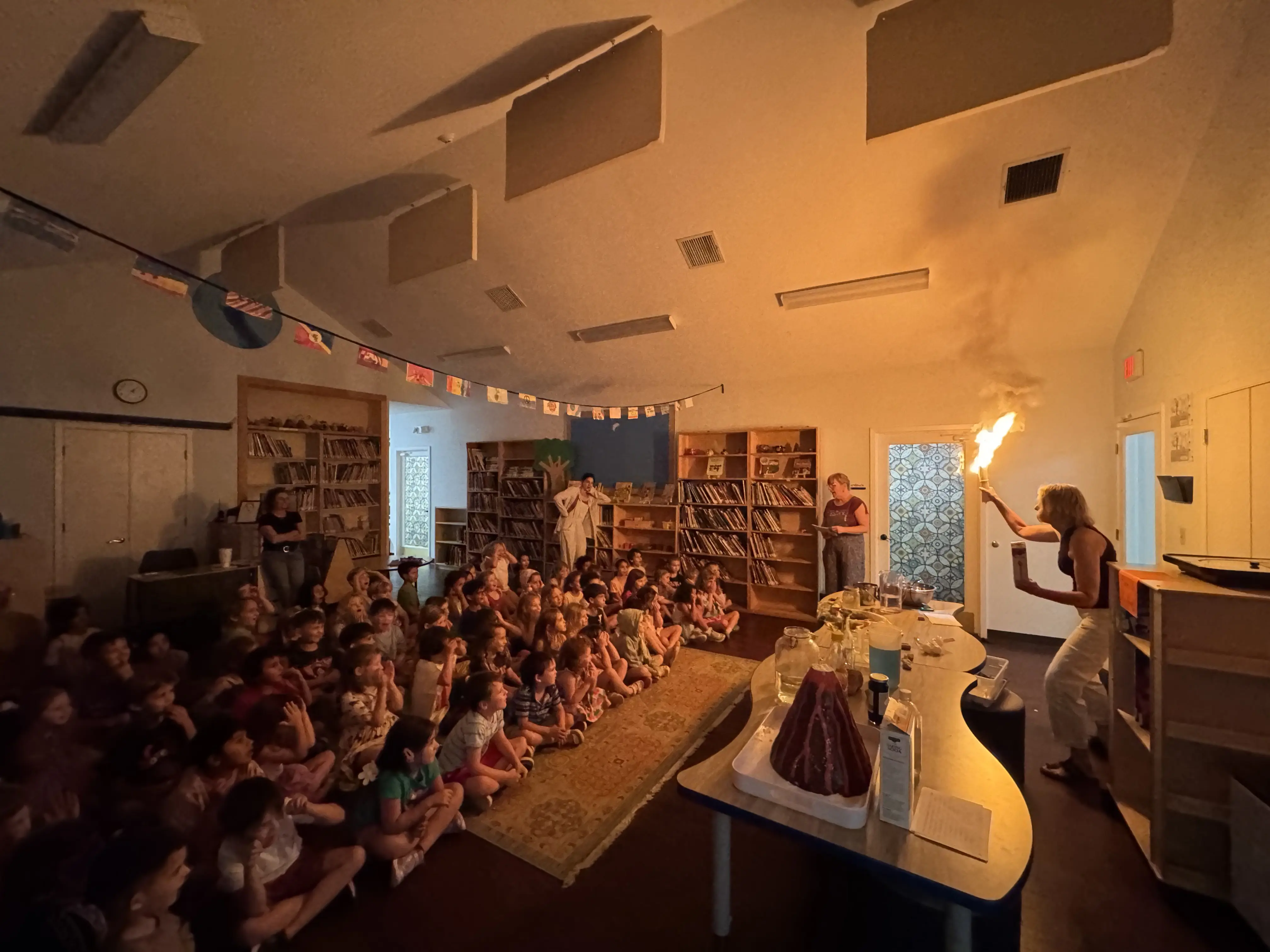 K–1 students sit gathered in a darkened classroom watching a guide demonstrate the Montessori First Great Lesson with a flame experiment during a presentation on the Story of the Universe.