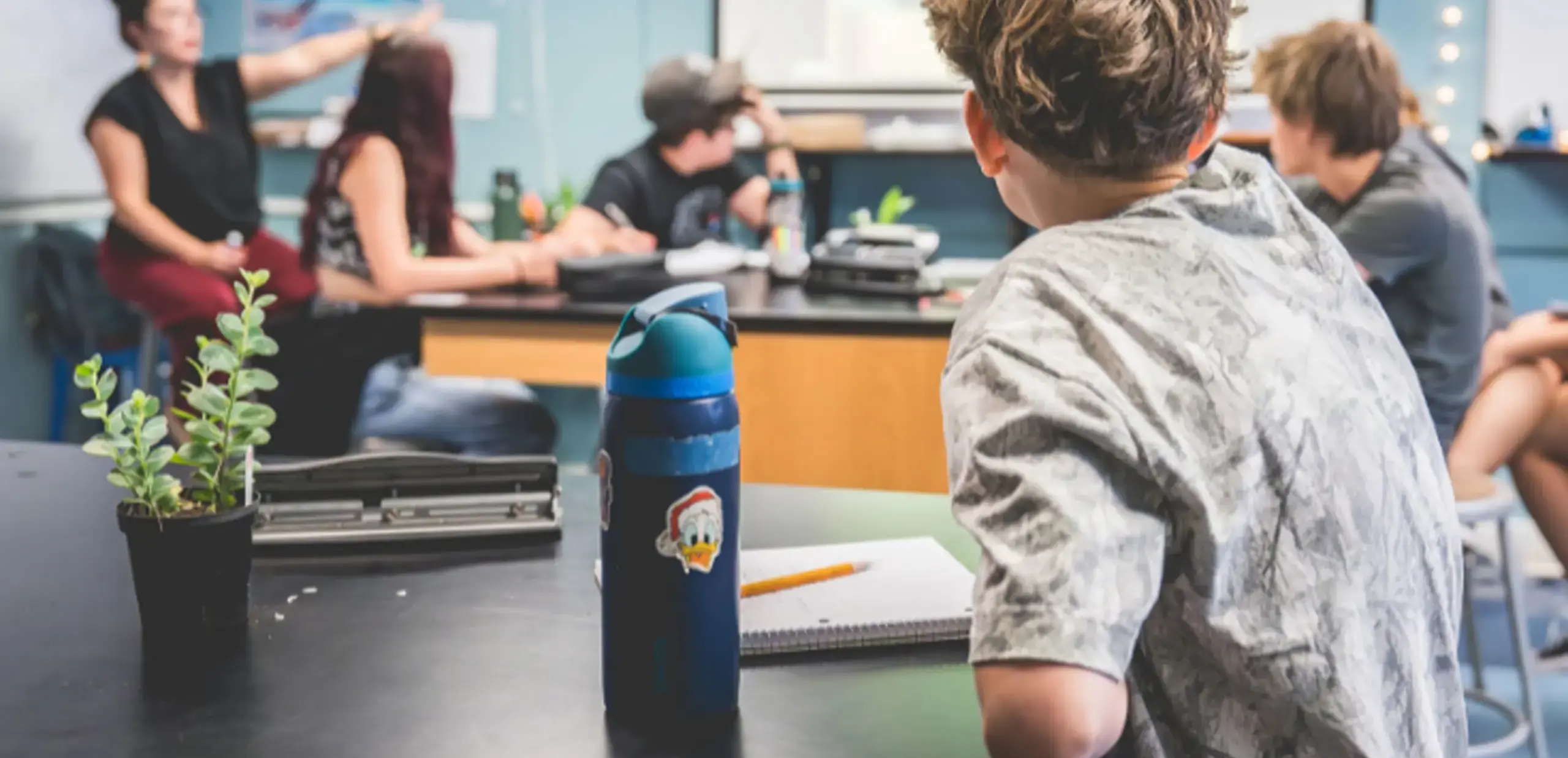 Middle school students in a science classroom, with a teacher gesturing toward a wall display while students work at lab tables.