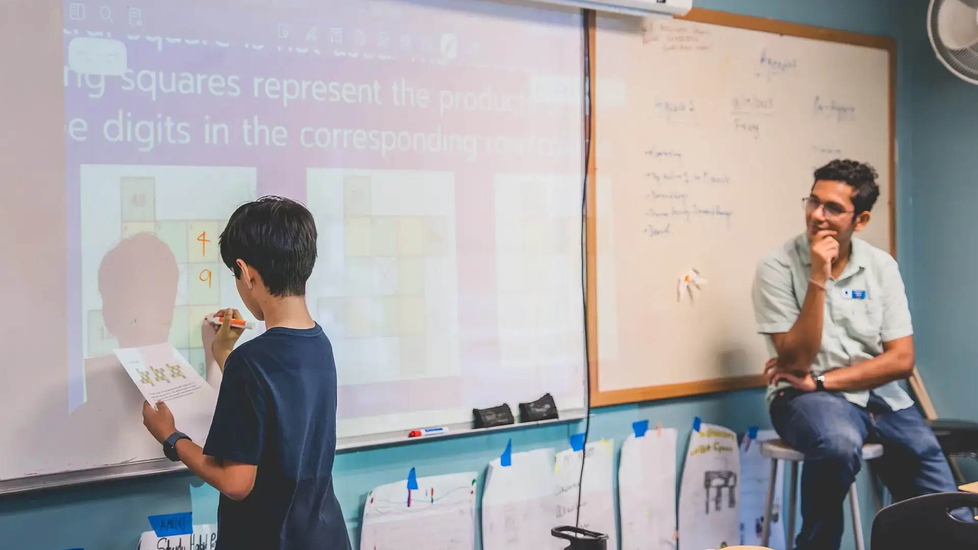 a middle school student solving a math problem on a whiteboard while a teacher observes.