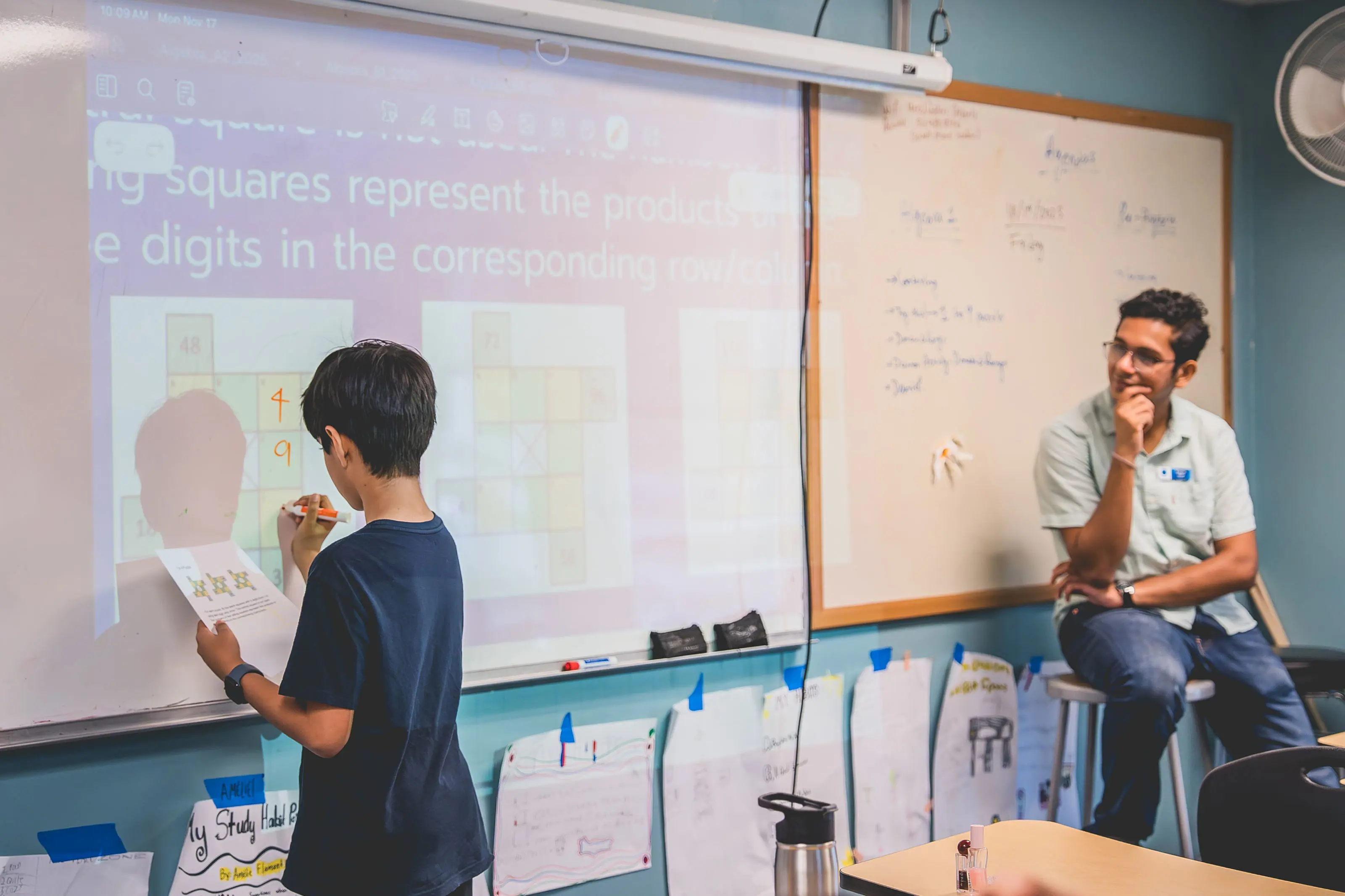 middle school student solving a problem on the whiteboard with a teacher looking on.