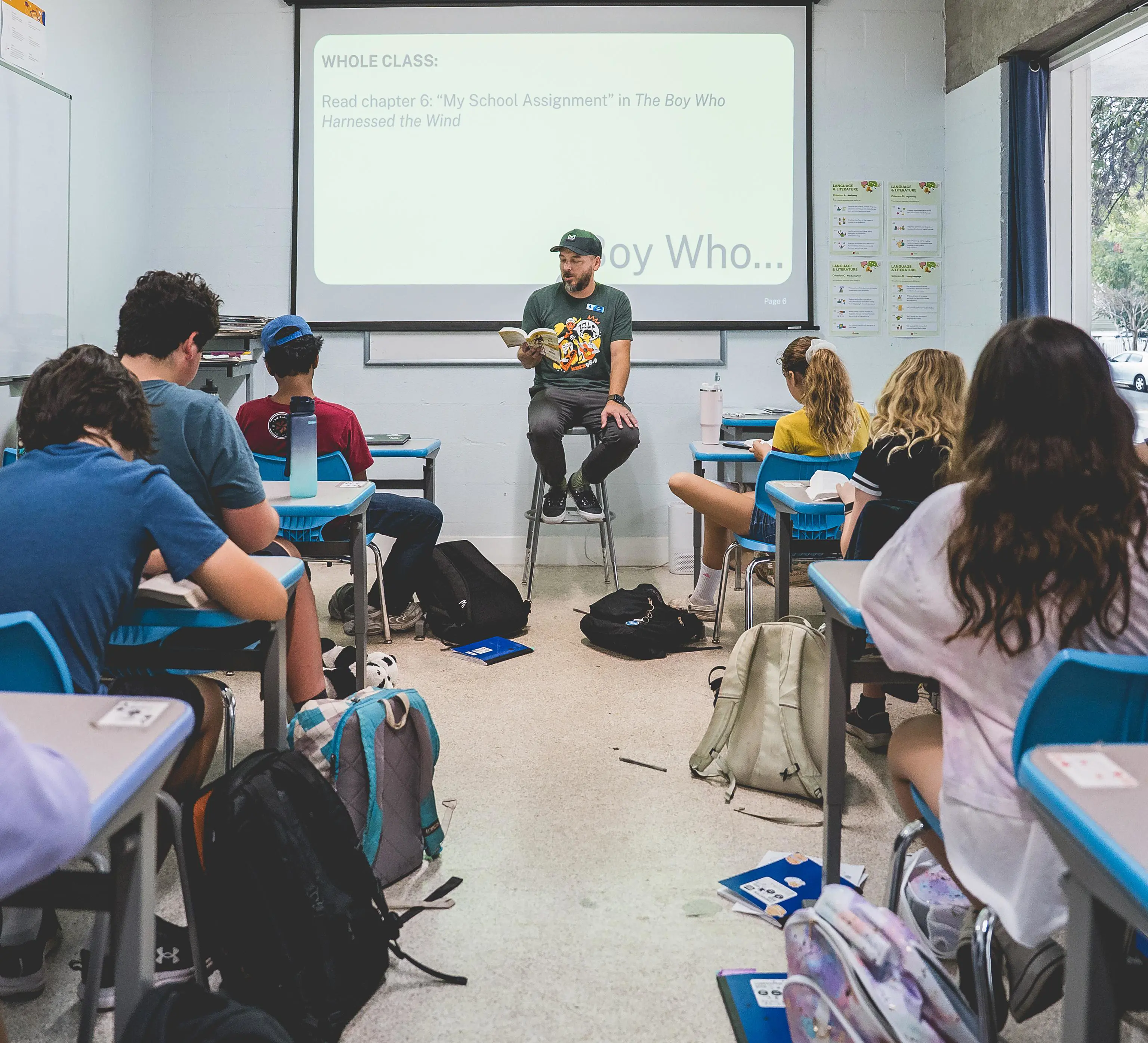 A teacher reads aloud to students seated at desks while a slide on "The Boy Who Harnessed the Wind" is projected behind him.