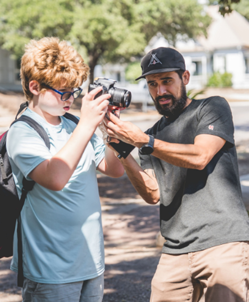 teacher wearing a black cap helps a student with glasses hold and look through a digital camera outdoors on a sunny day.