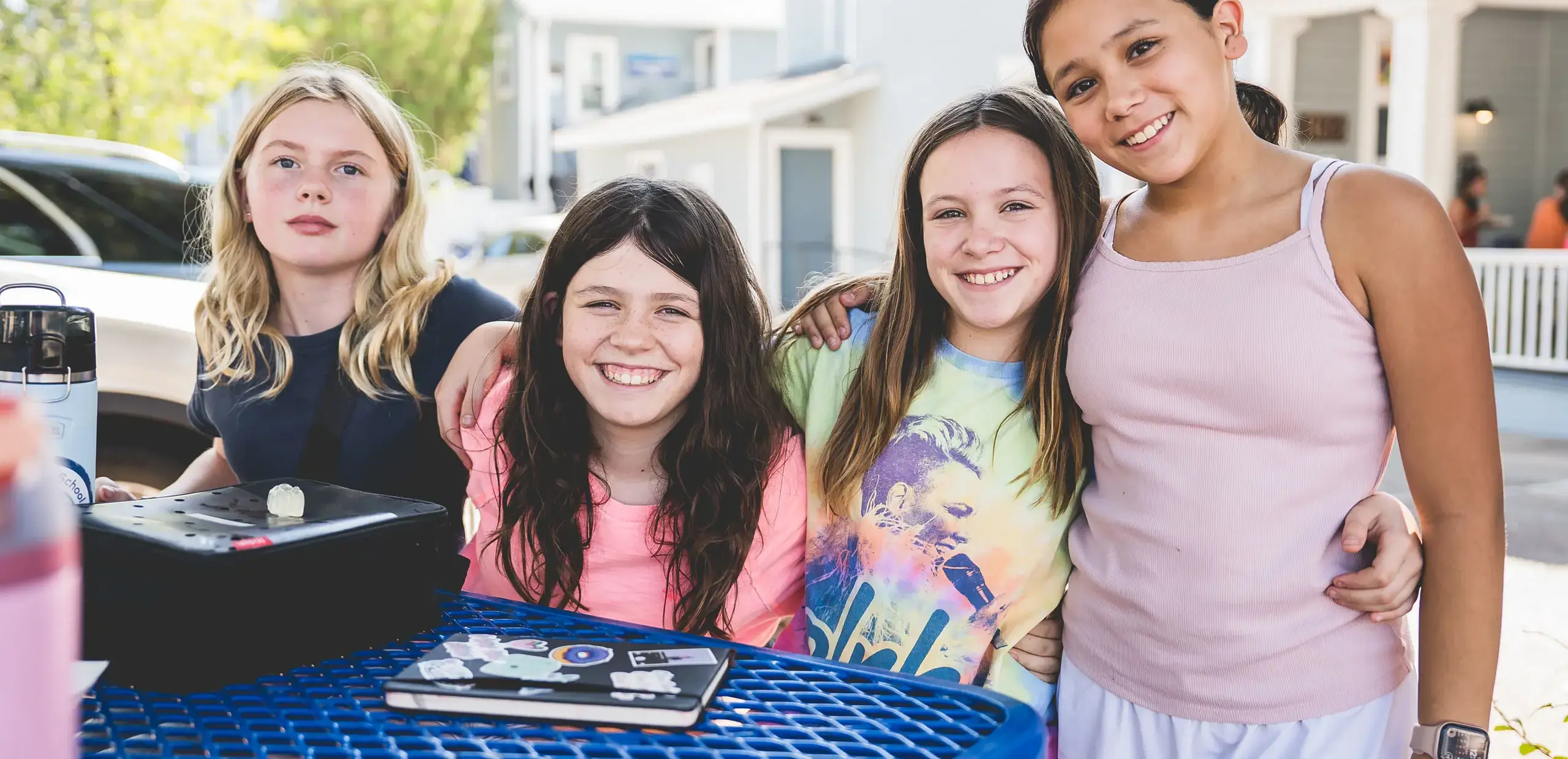 Four middle school girls smiling with arms around each other at an outdoor picnic table.