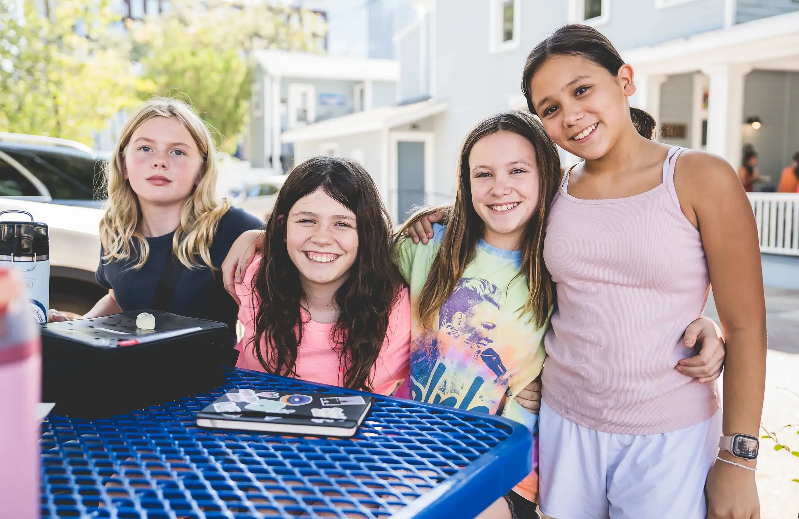 Four middle school students smiling and sitting together at a blue picnic table outside on campus