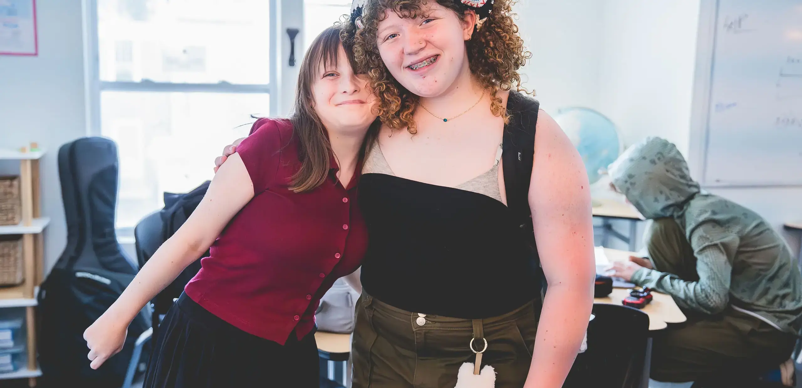 two middle school students smiling with an arm around each other in the classroom.
