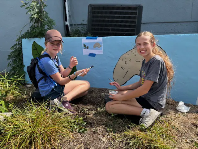 Middle school students painting mural together outdoors on campus.