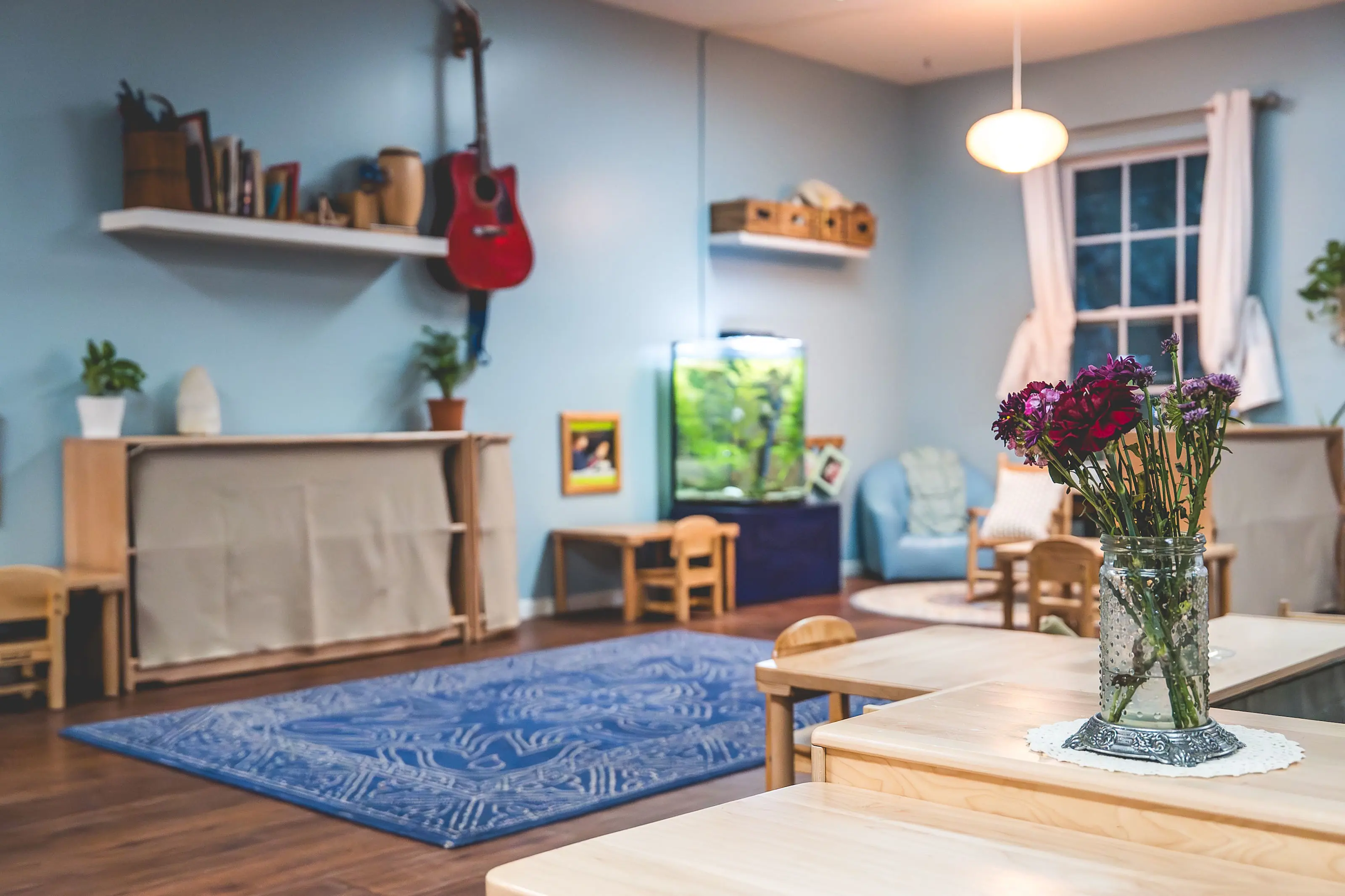 A calm Montessori classroom with wooden tables, a vase of flowers, learning shelves, and a cozy reading area.