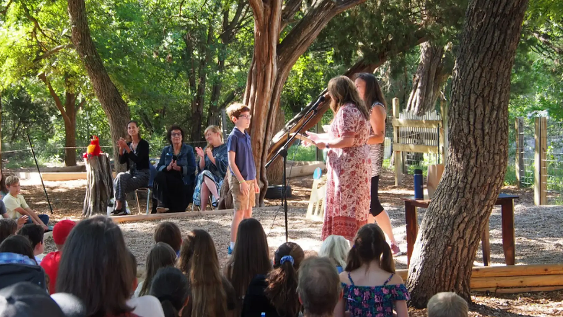 A speaker addressing an audience seated outdoors under trees during a ceremony.