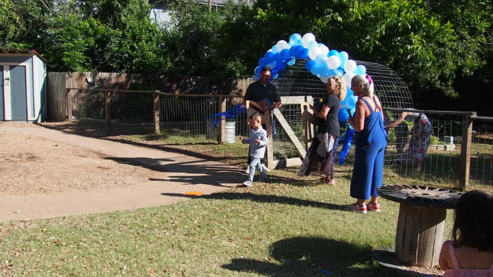 A graduate walking outdoors past a blue balloon arch during a celebration.